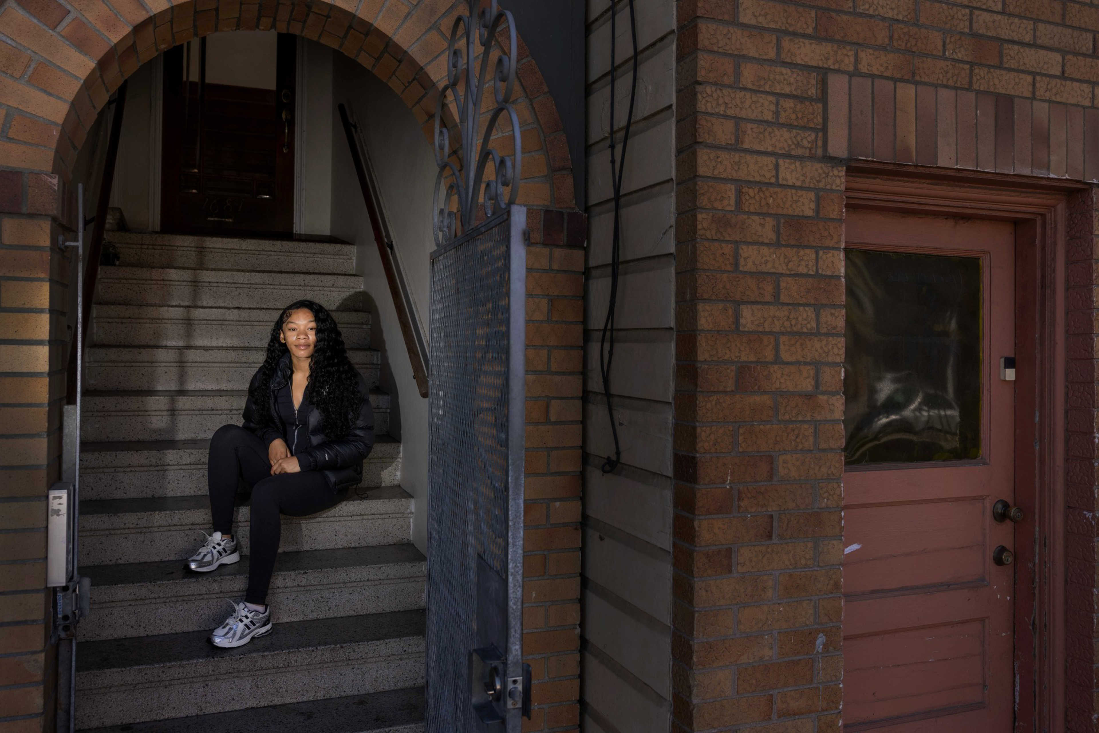A woman with long curly hair wearing black clothes and white sneakers sits on a stone staircase under an arched brick entrance with a metal gate.