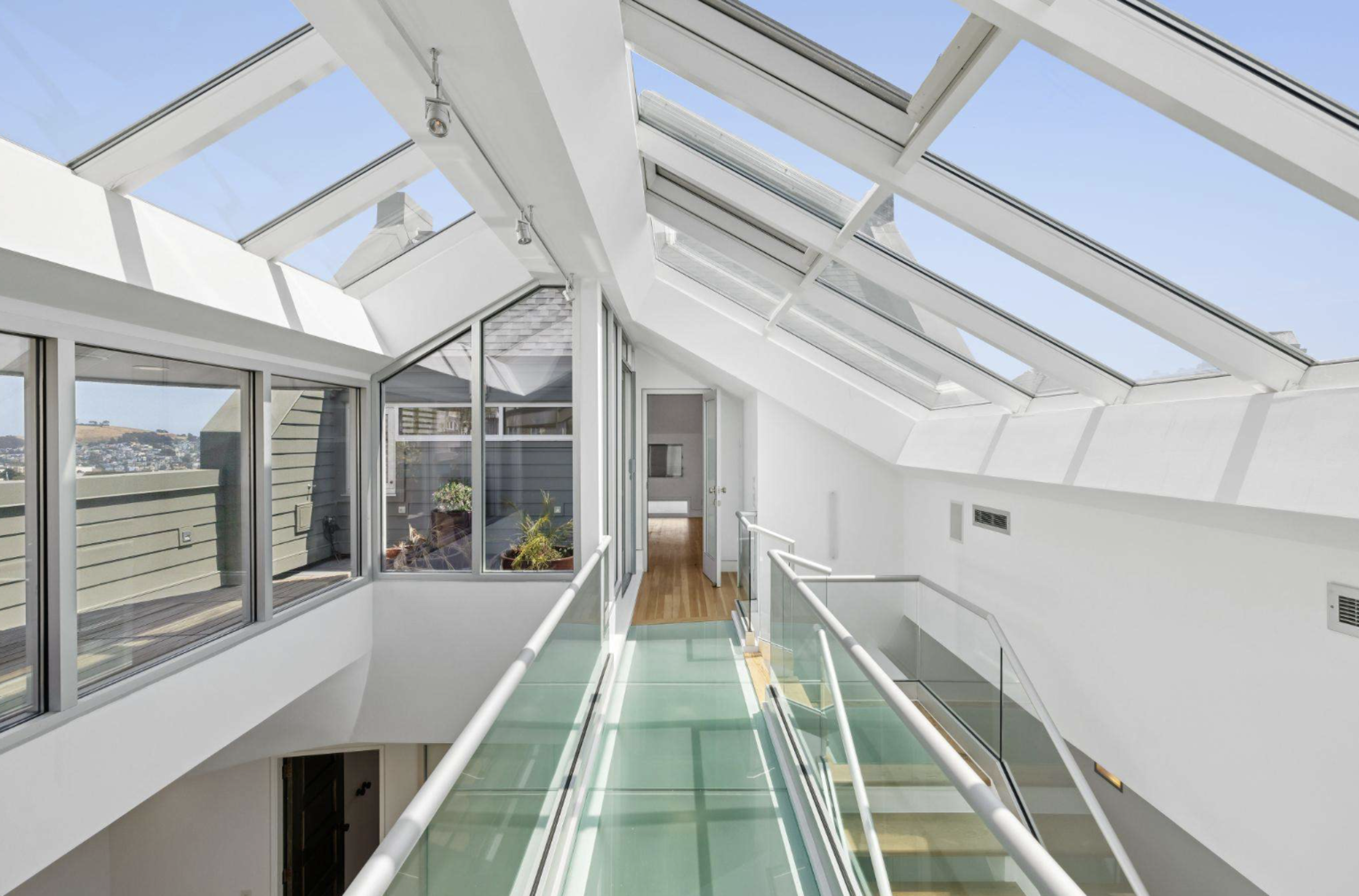 A bright hallway with glass floors and railings is illuminated by skylights and large windows, featuring modern white walls and a wooden floor at the far end.