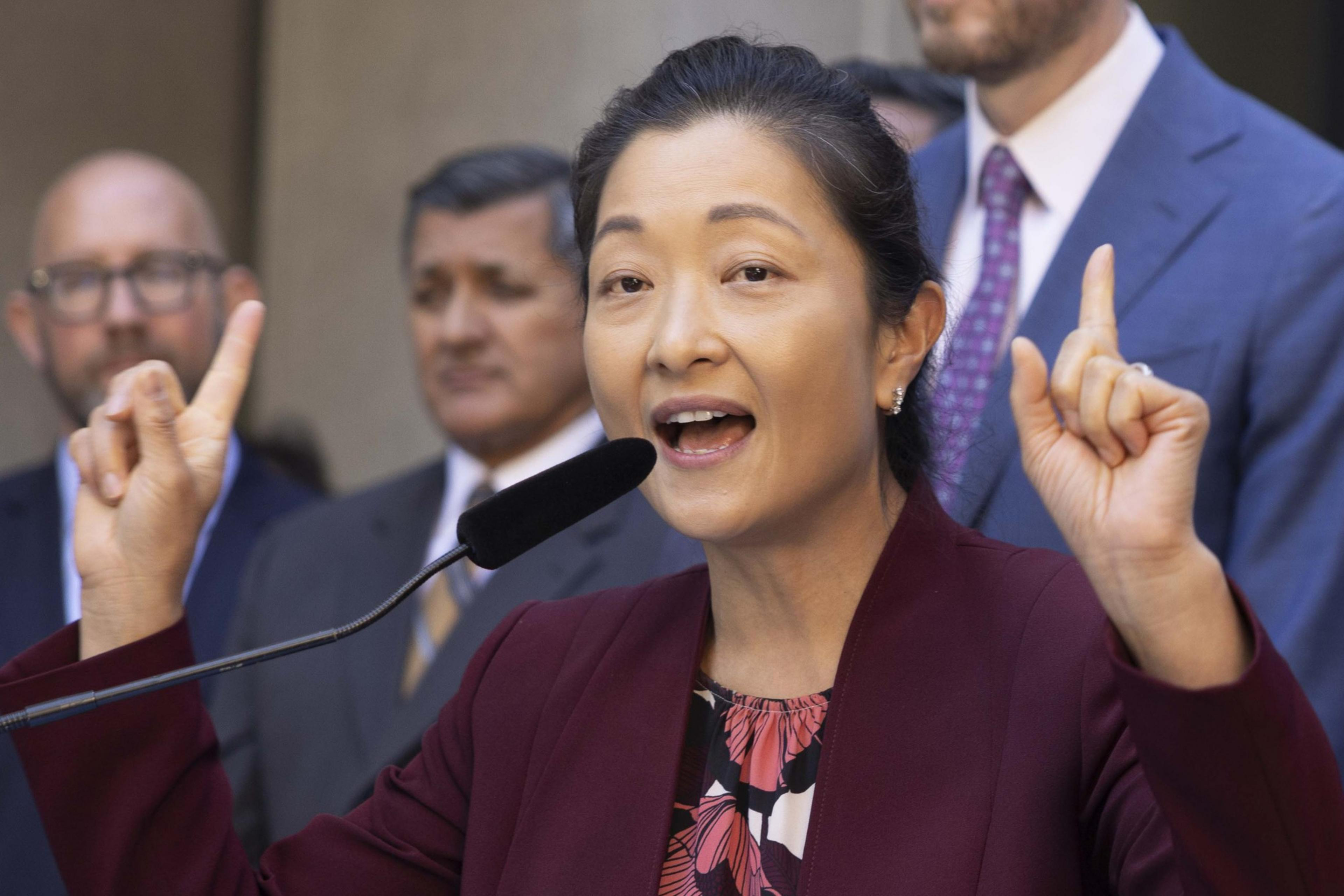 A woman speaks passionately into a microphone with both index fingers raised, while three men in suits stand behind her.