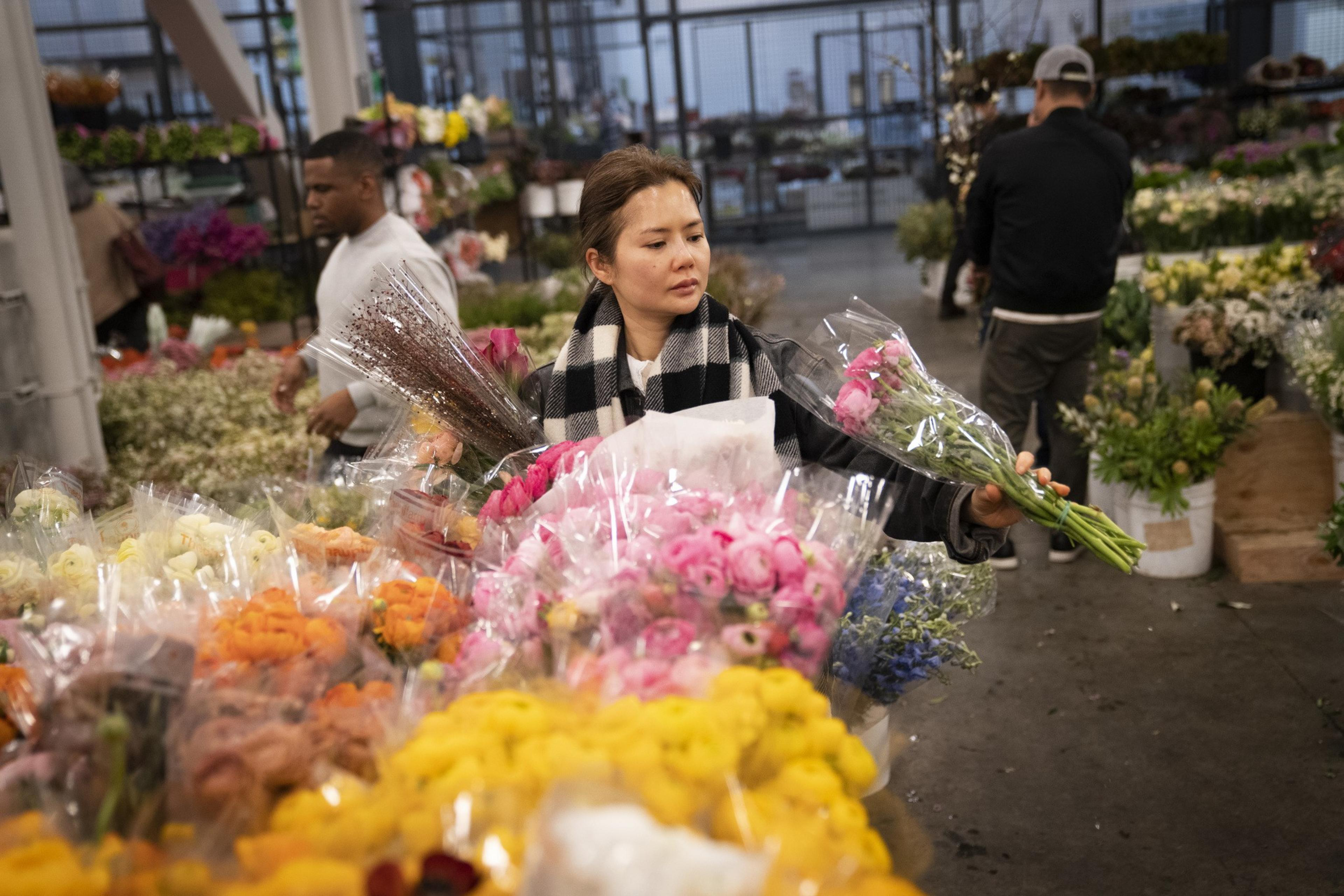 A woman in a scarf selects pink flowers in a bustling flower market, surrounded by colorful bouquets and bustling shoppers in the background.