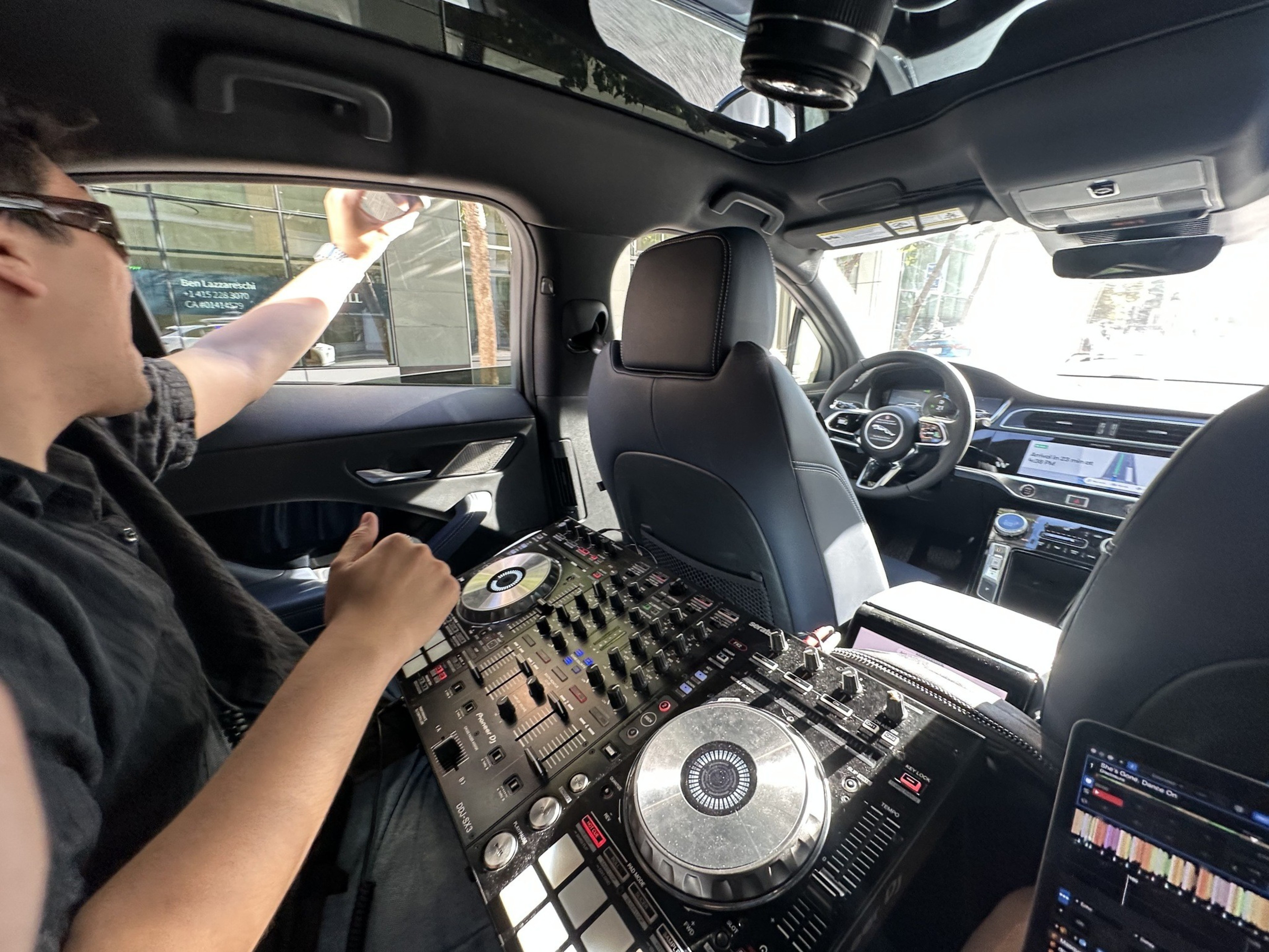 A person is sitting in the backseat of a car, using DJ equipment placed between the seats. They're facing forward, and sunlight streams in from the windows.