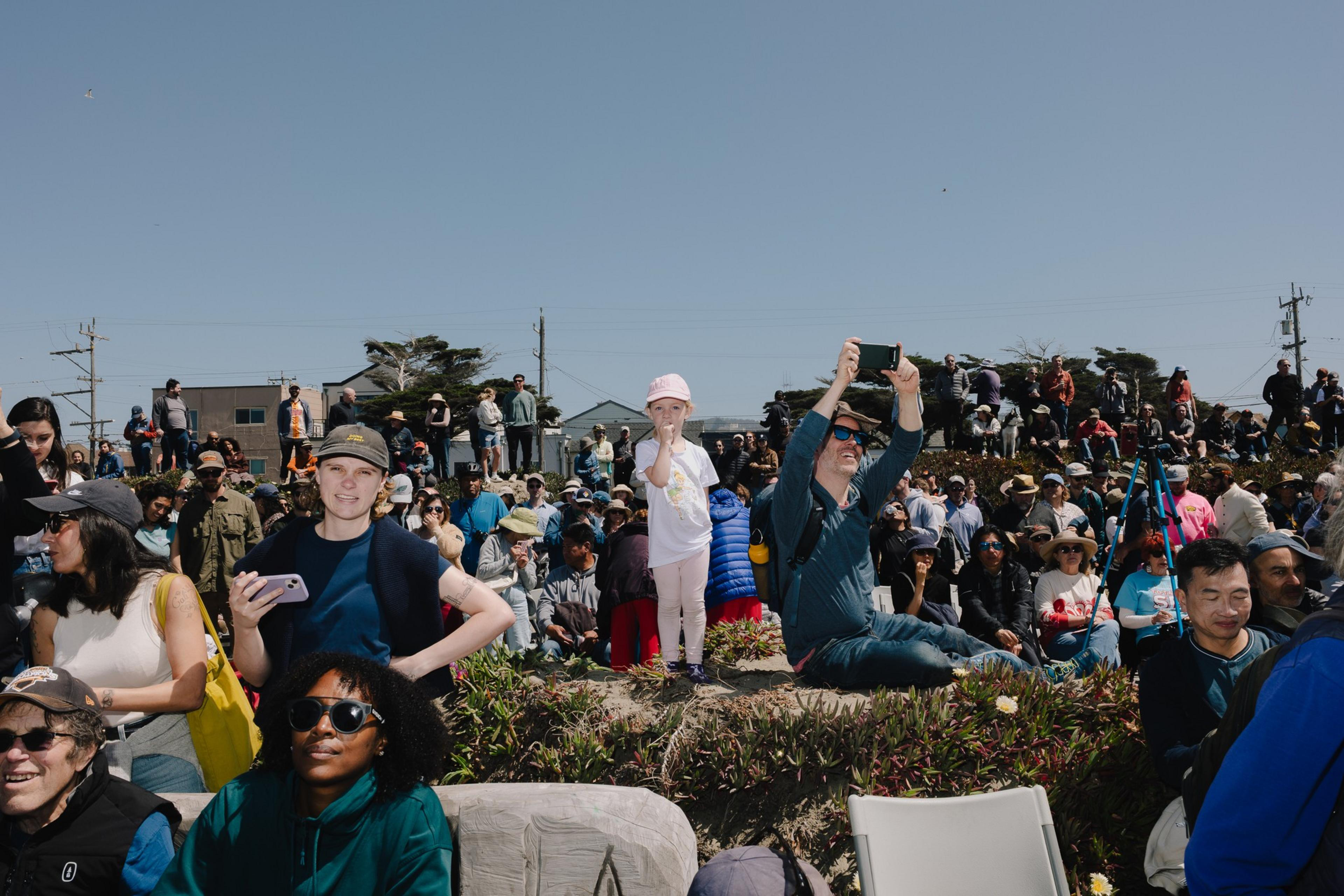 A large crowd of people is gathered outdoors under a clear blue sky. A child stands on a small hill, surrounded by adults, some taking photos.