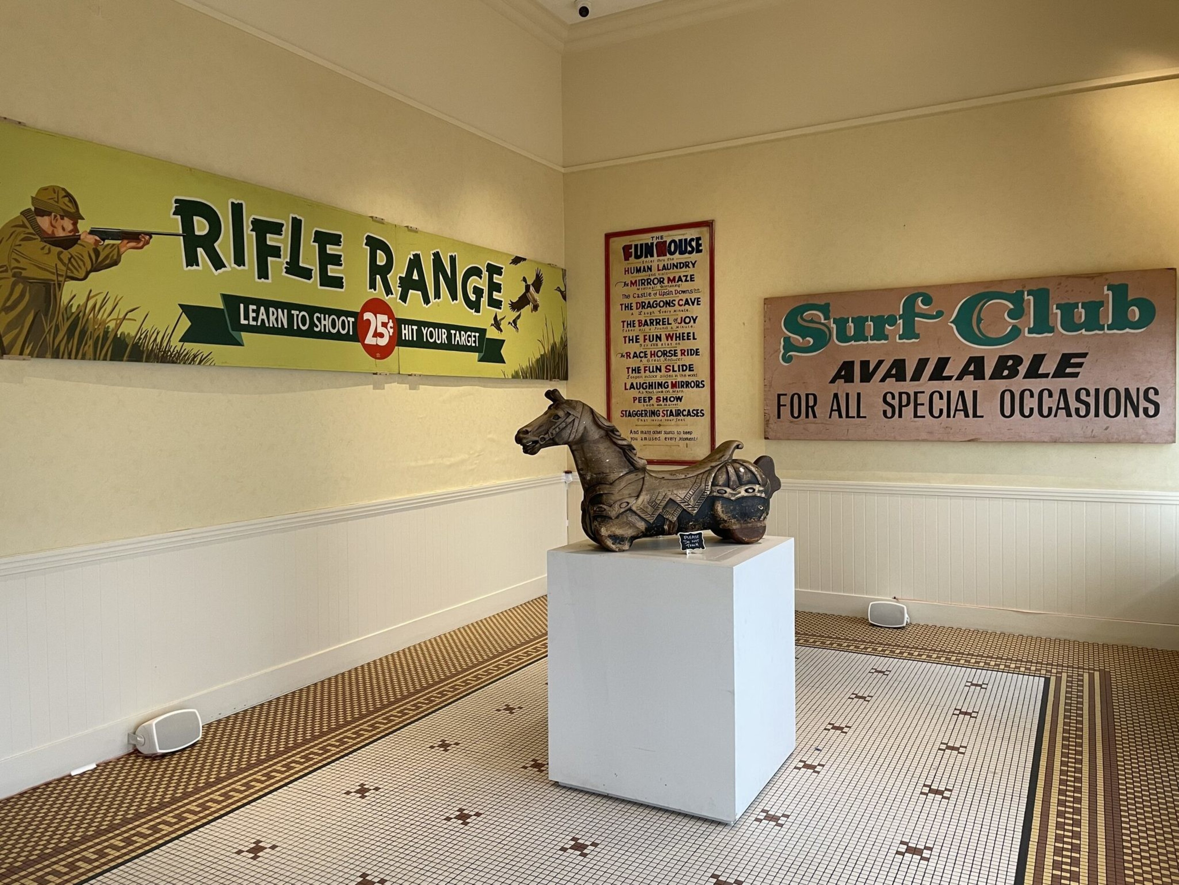 An indoor corner with vintage signs for a rifle range, a surf club, and a funhouse, featuring a carved wooden horse statue on a white pedestal.
