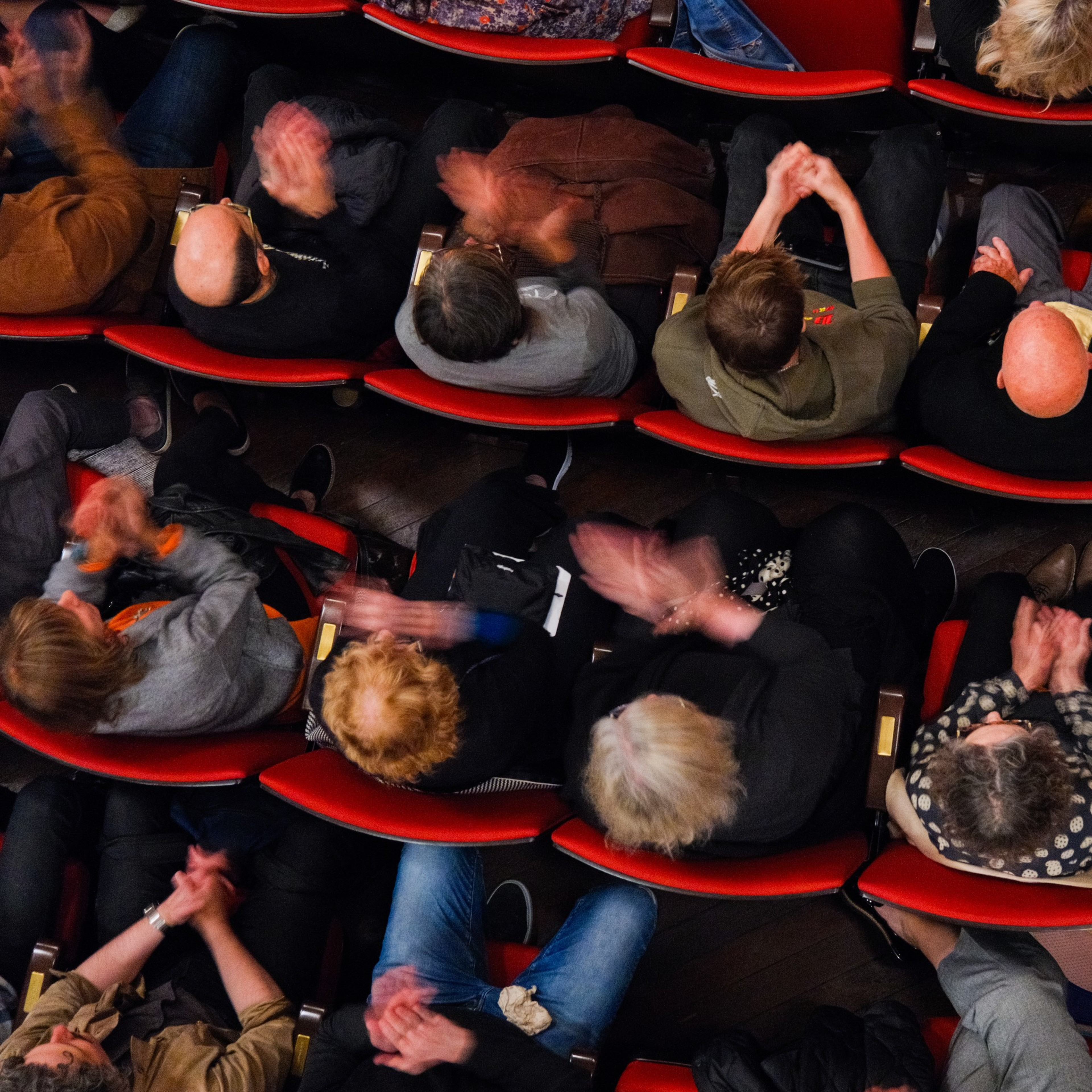 An overhead view of an audience seated in red theater chairs, clapping enthusiastically. The image captures a mix of people, some mid-clap, creating a blur of motion.