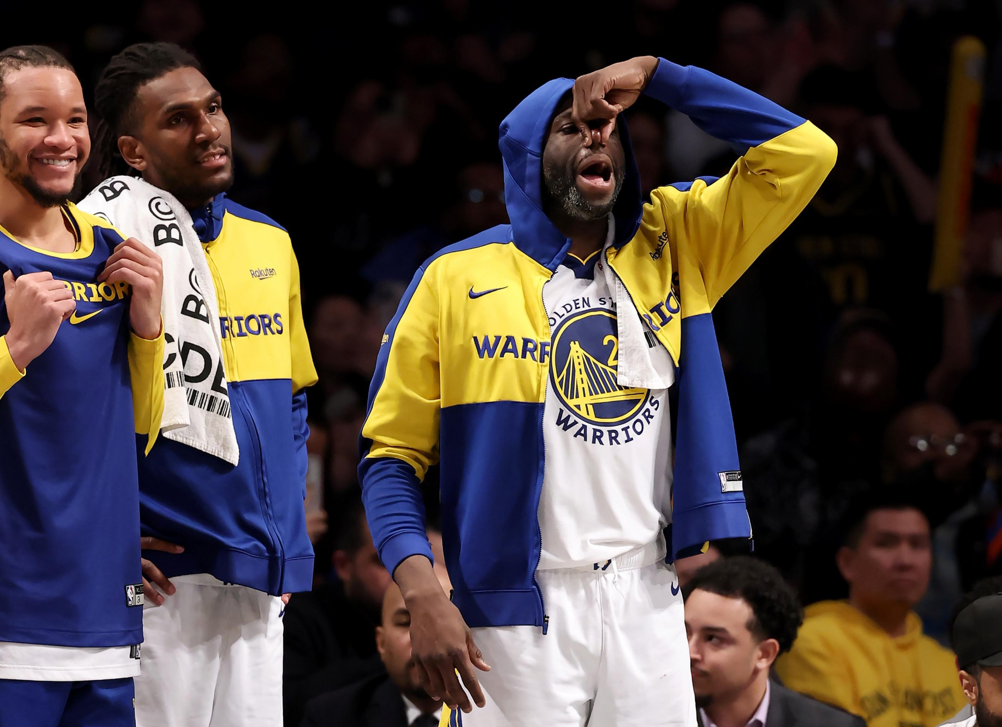 Three basketball players in Warriors gear are on the sideline. One is pinching his nose in a humorous gesture, while the others are smiling and watching.