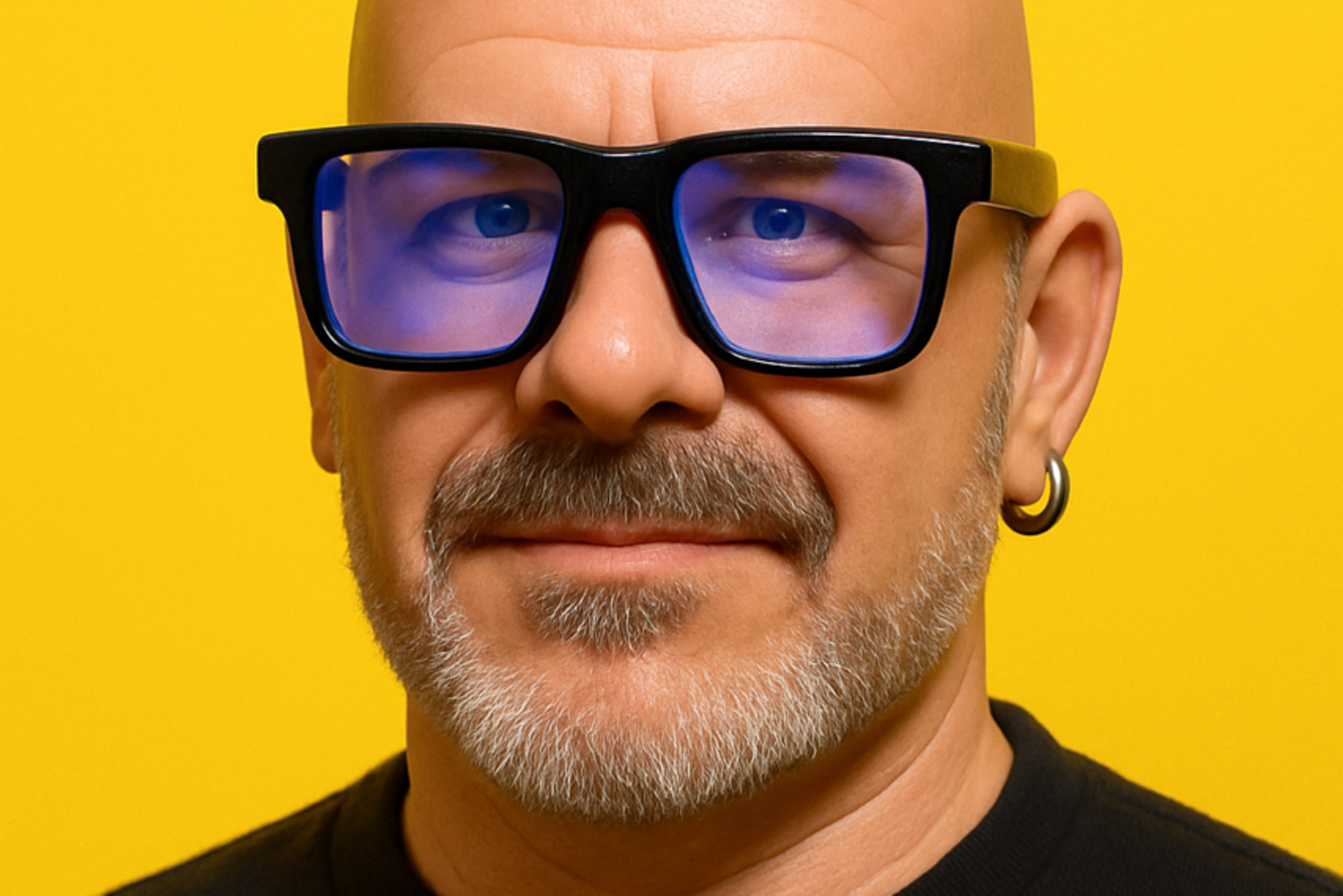 A bald man with a gray beard wears large blue-tinted glasses, a black shirt with "artificial" printed on it, and a small hoop earring, against a yellow background.
