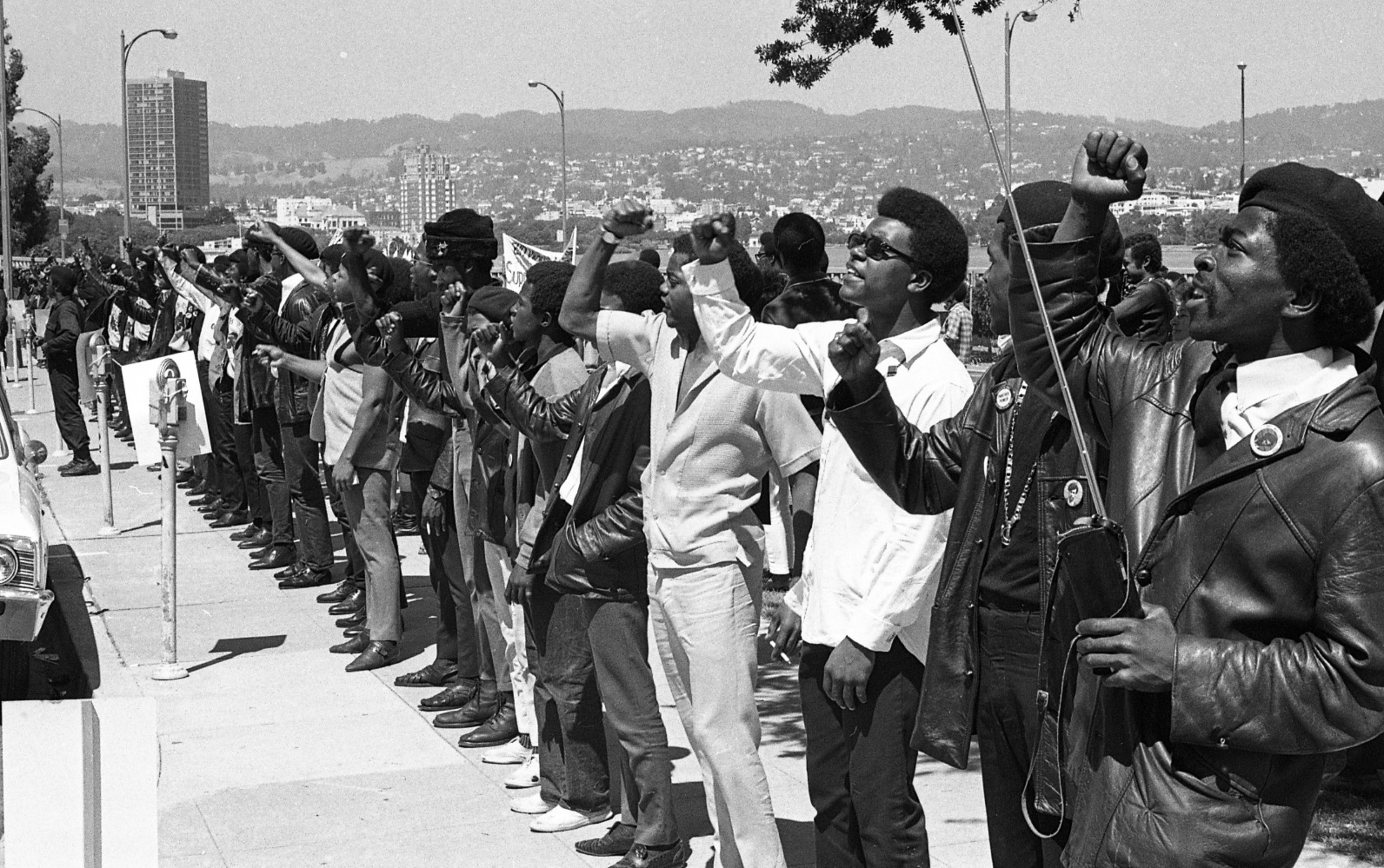 A line of men, many wearing leather jackets and sunglasses, stand with raised fists in a Black Power salute along a sidewalk with a cityscape in the background.