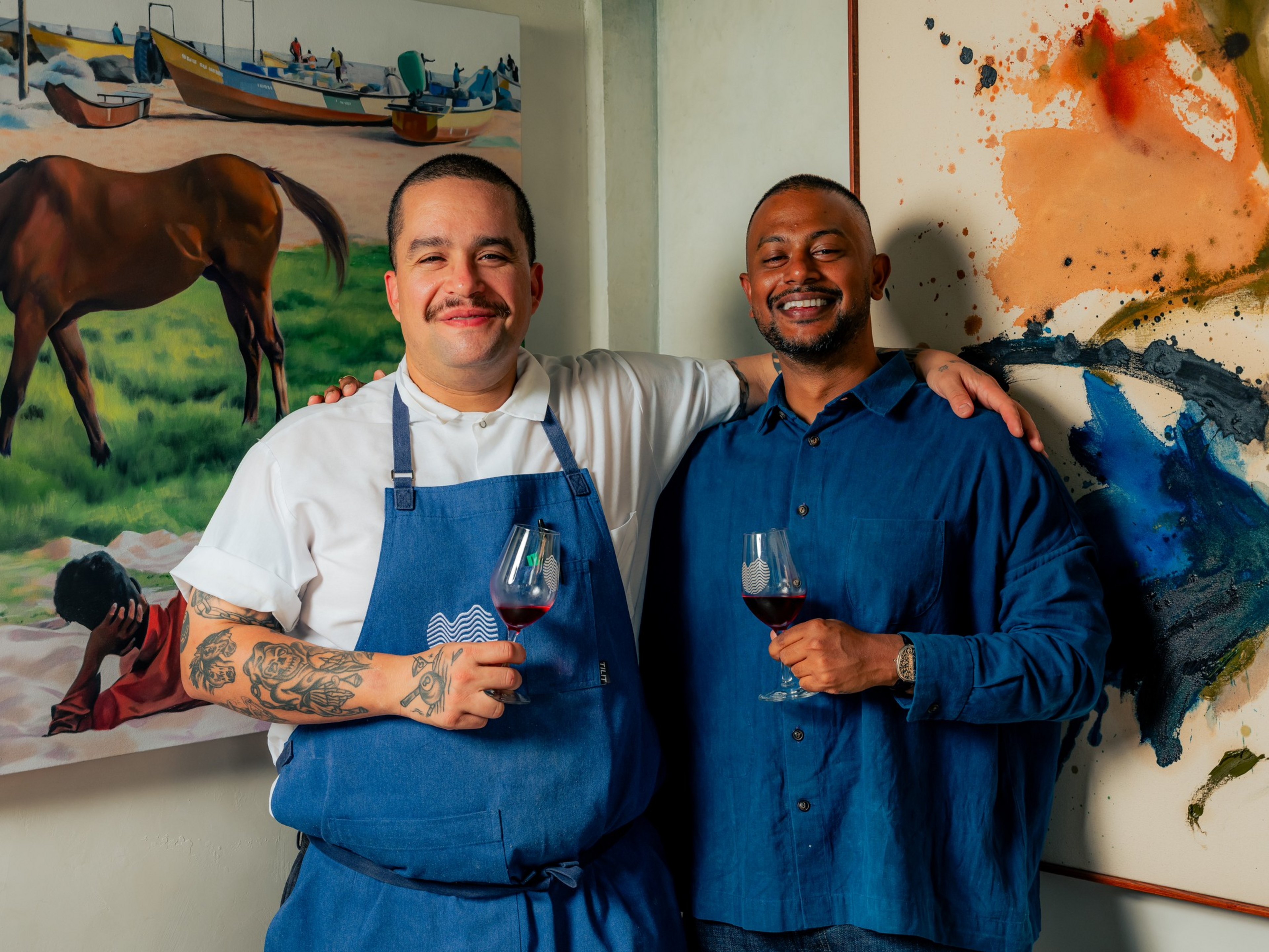 Two men smiling with arms around each other, holding glasses of red wine, one wearing a white shirt and blue apron, the other in a blue shirt.