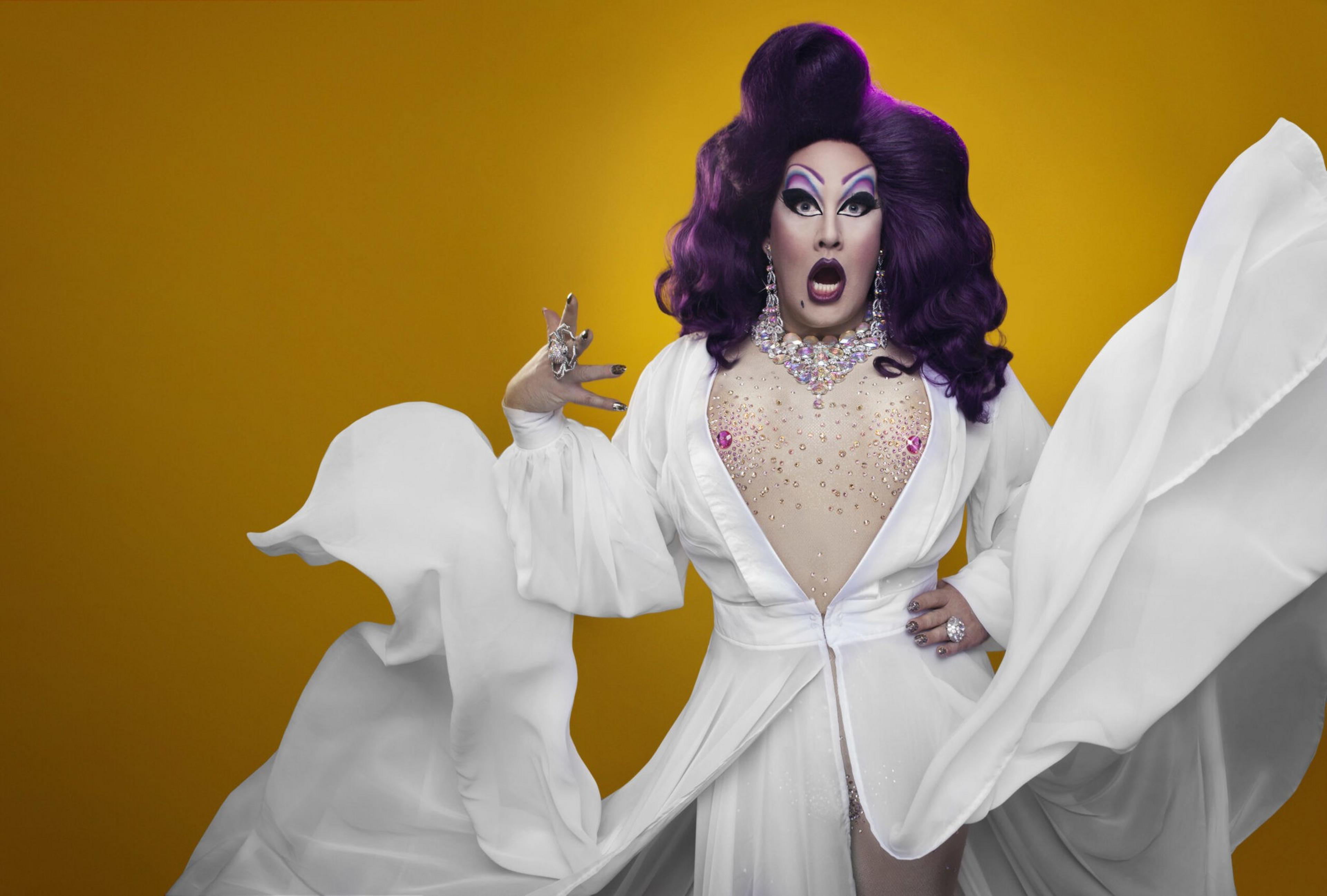 A person with dramatic purple hair and bold makeup wears a flowing white gown with crystal embellishments and an open front, posing against a yellow background.