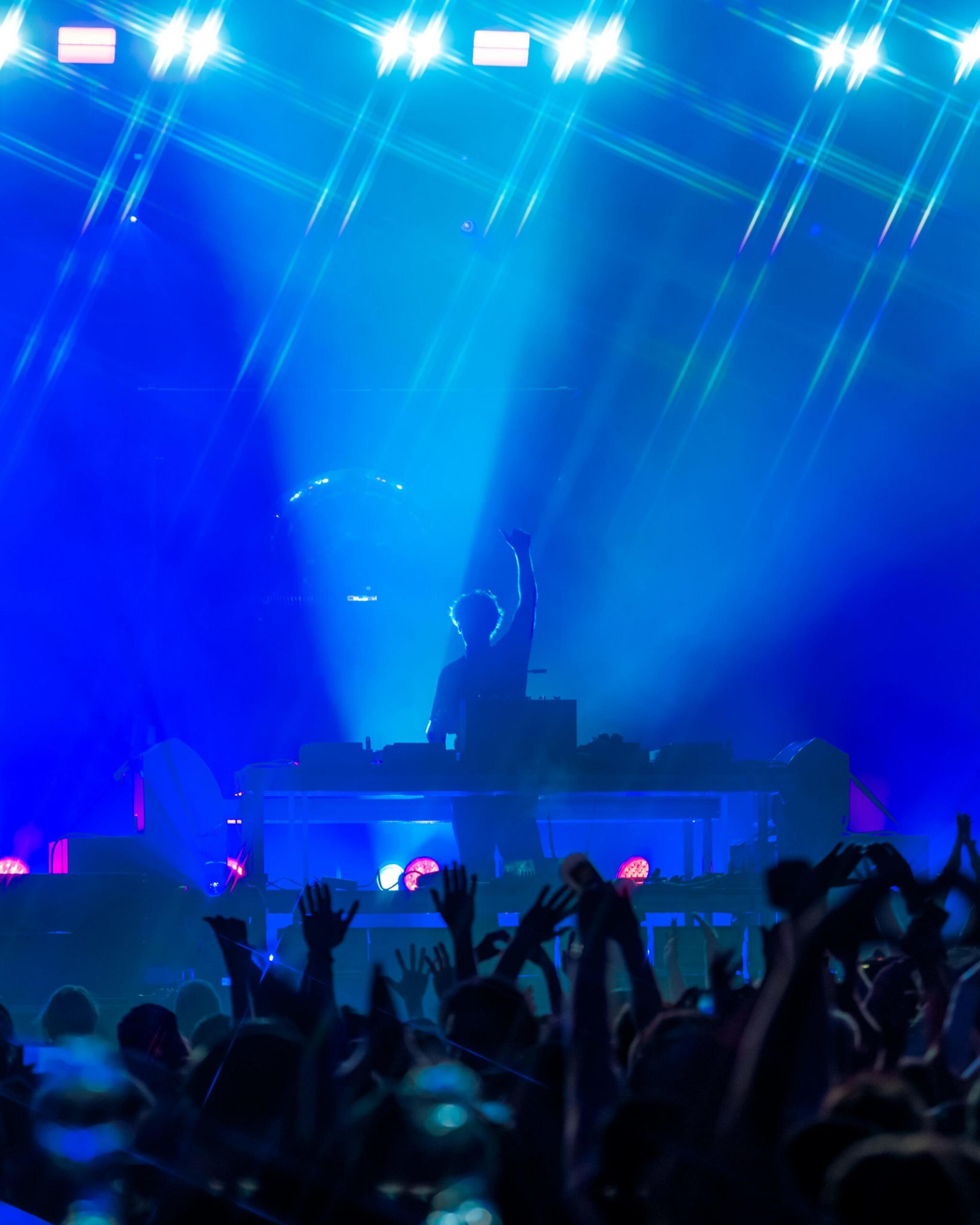A DJ stands behind a table with equipment, raising one hand, illuminated by blue stage lights, as a crowd with raised hands enjoys the music.