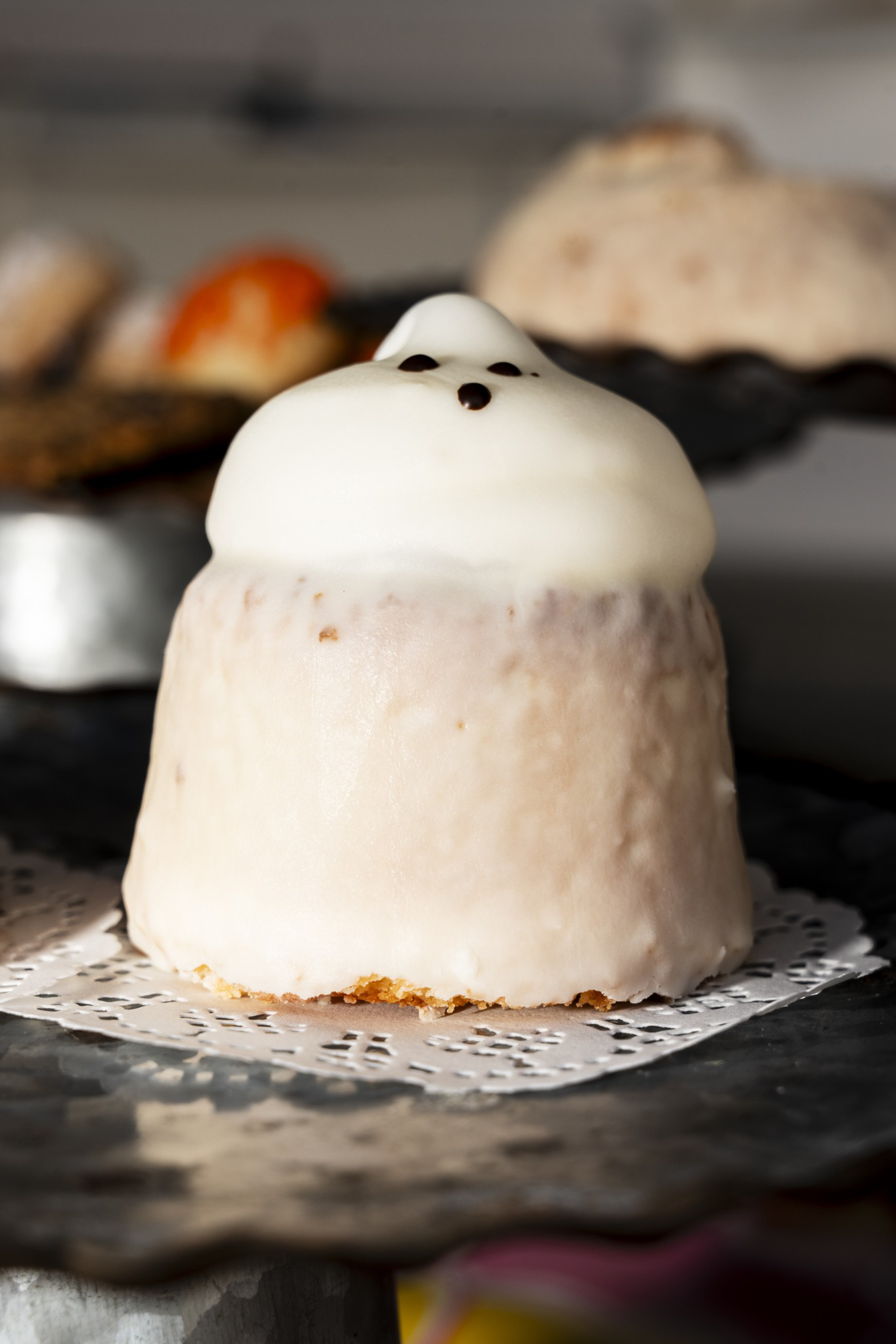 A small dessert covered in white icing with three tiny dark dots on top, resembling a ghost, sits on a doily on a dark tray.
