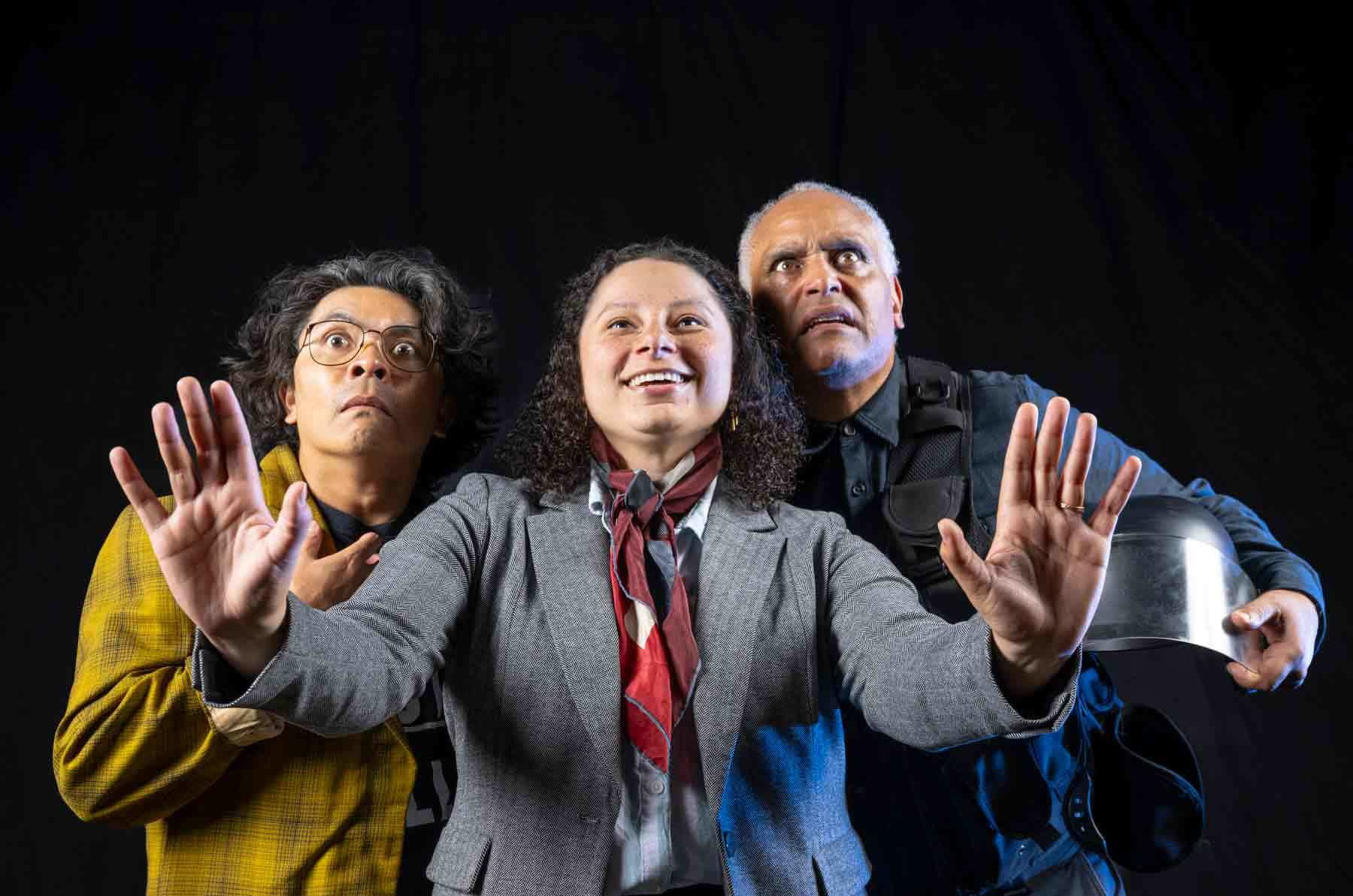Three people stand close together against a dark background. The person in the middle smiles with hands raised, while the others look surprised, with one holding a helmet.