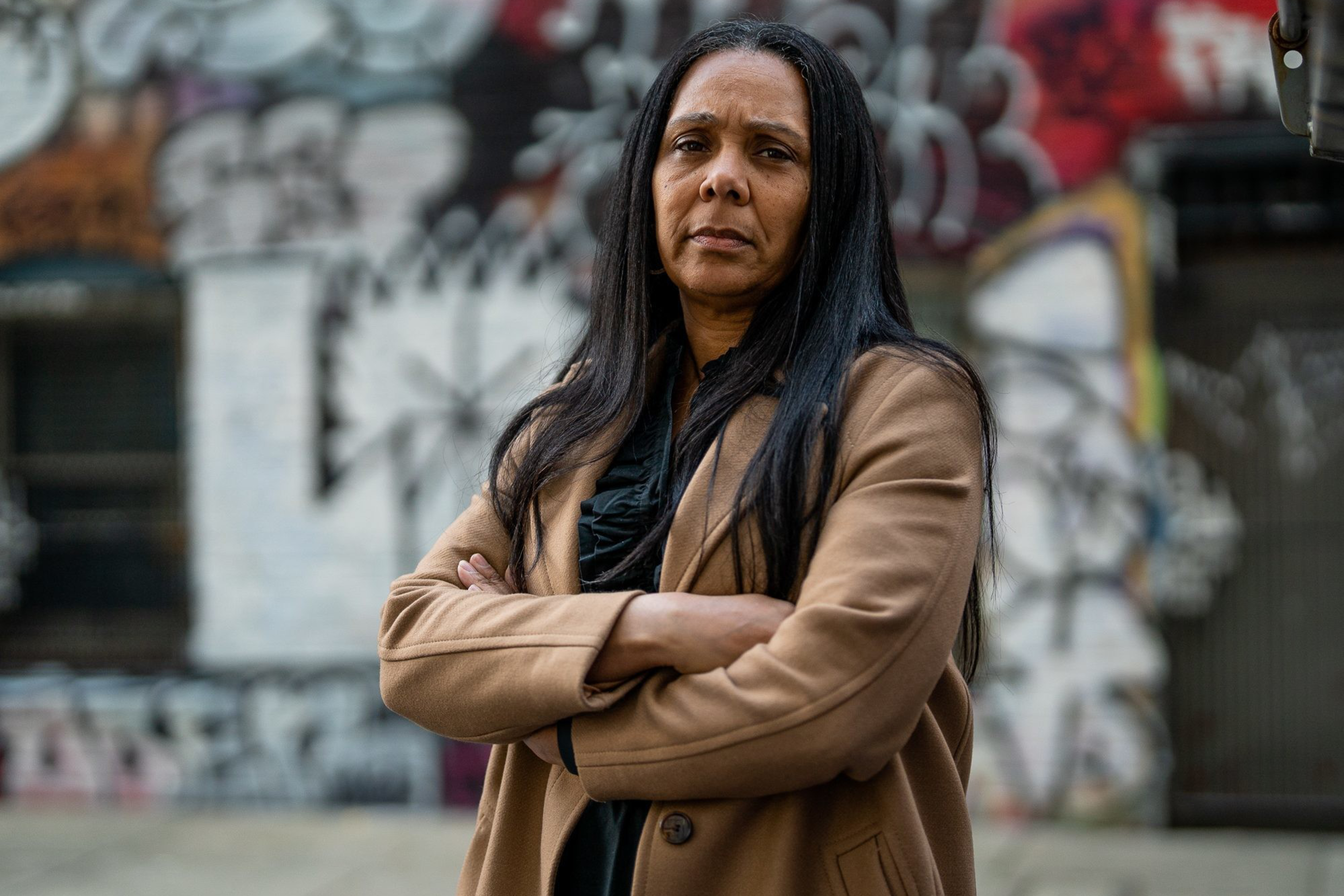A woman with long dark hair, wearing a tan coat, stands with arms crossed in front of a graffiti-covered wall on an urban street. She has a serious expression.