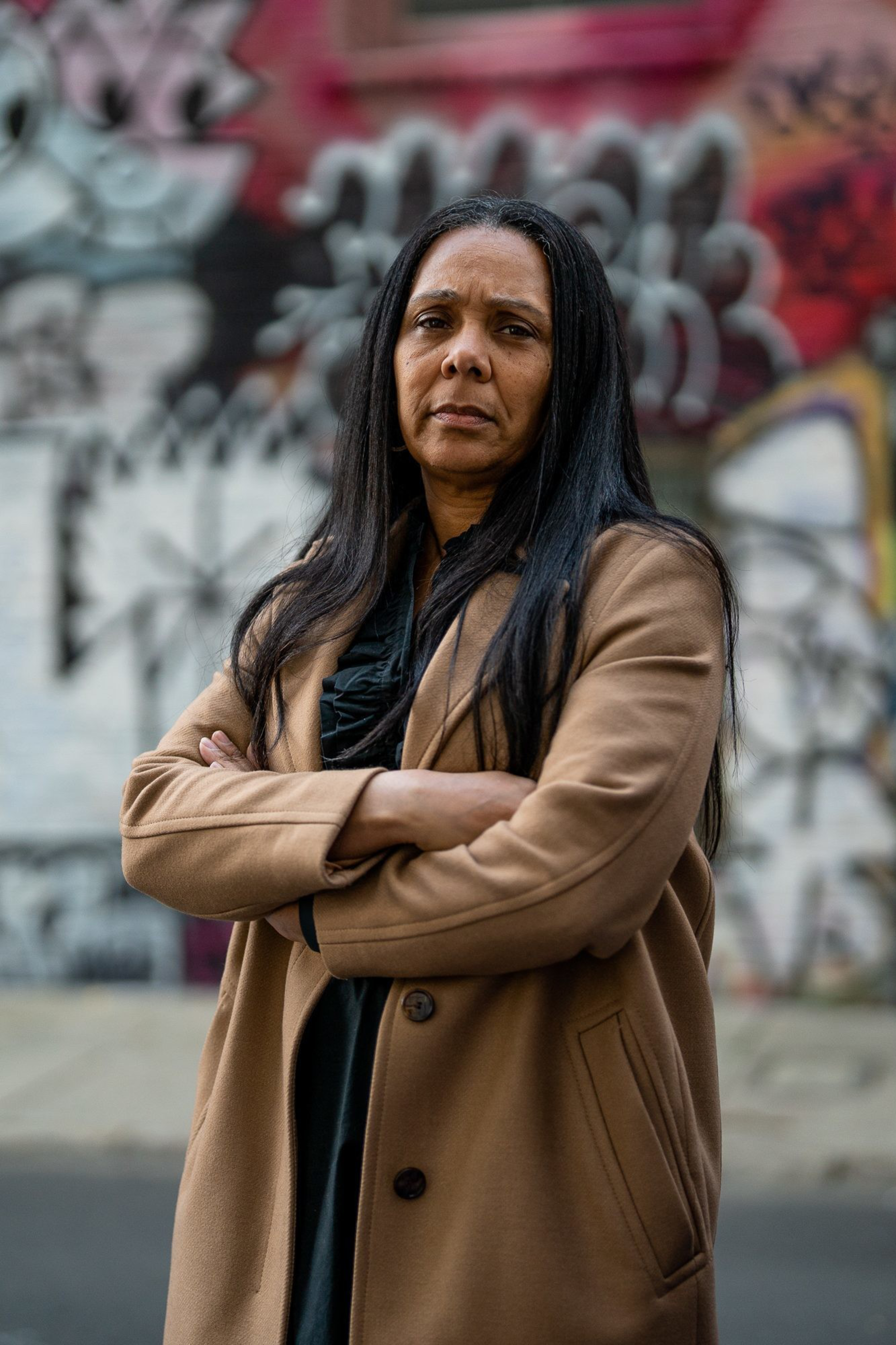 A woman with long dark hair, wearing a tan coat, stands with arms crossed in front of a graffiti-covered wall on an urban street. She has a serious expression.