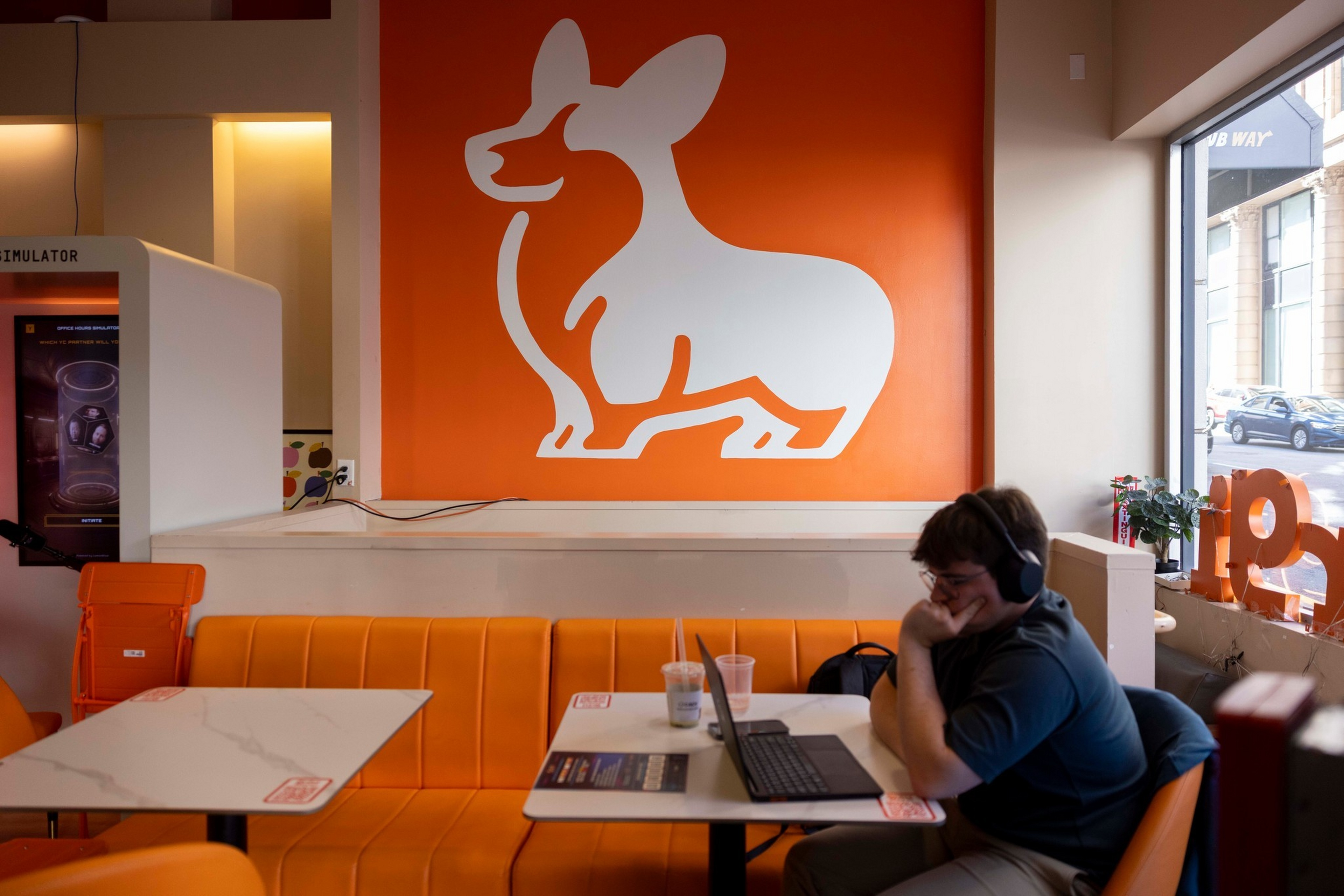 A person wearing headphones works on a laptop while seated at an orange booth beneath a large white corgi dog silhouette on an orange wall.