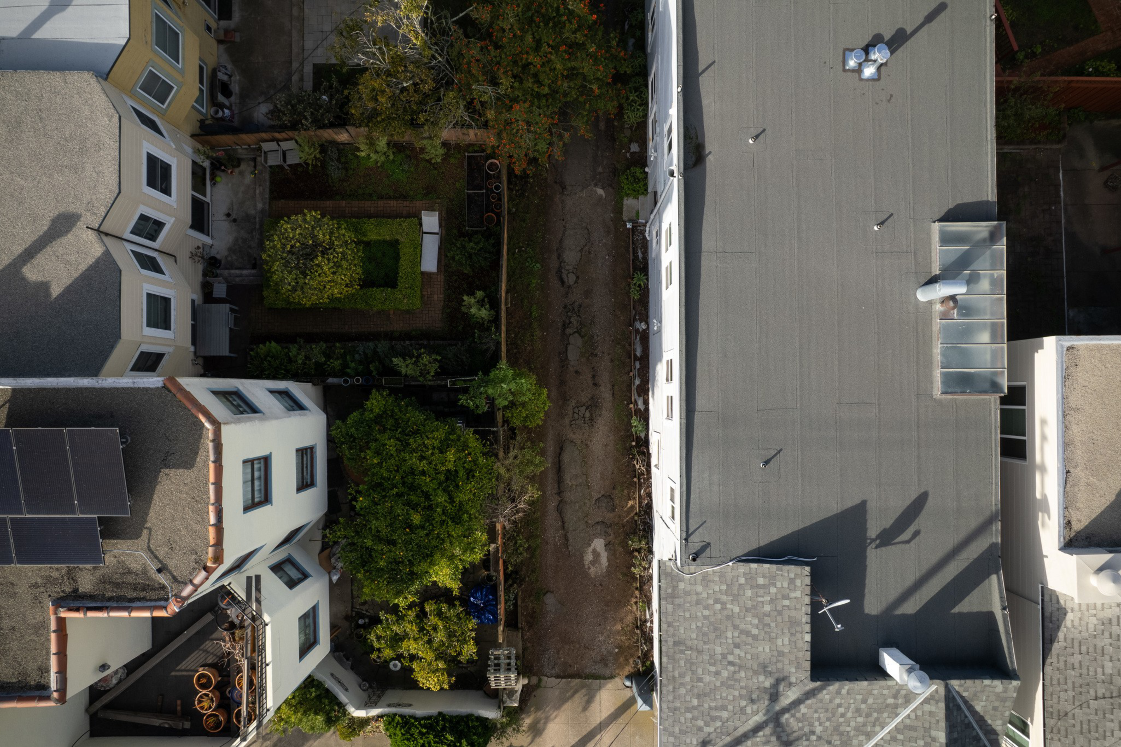 Two buildings border a narrow dirt path lined with green trees and small garden plots, one roof featuring solar panels.