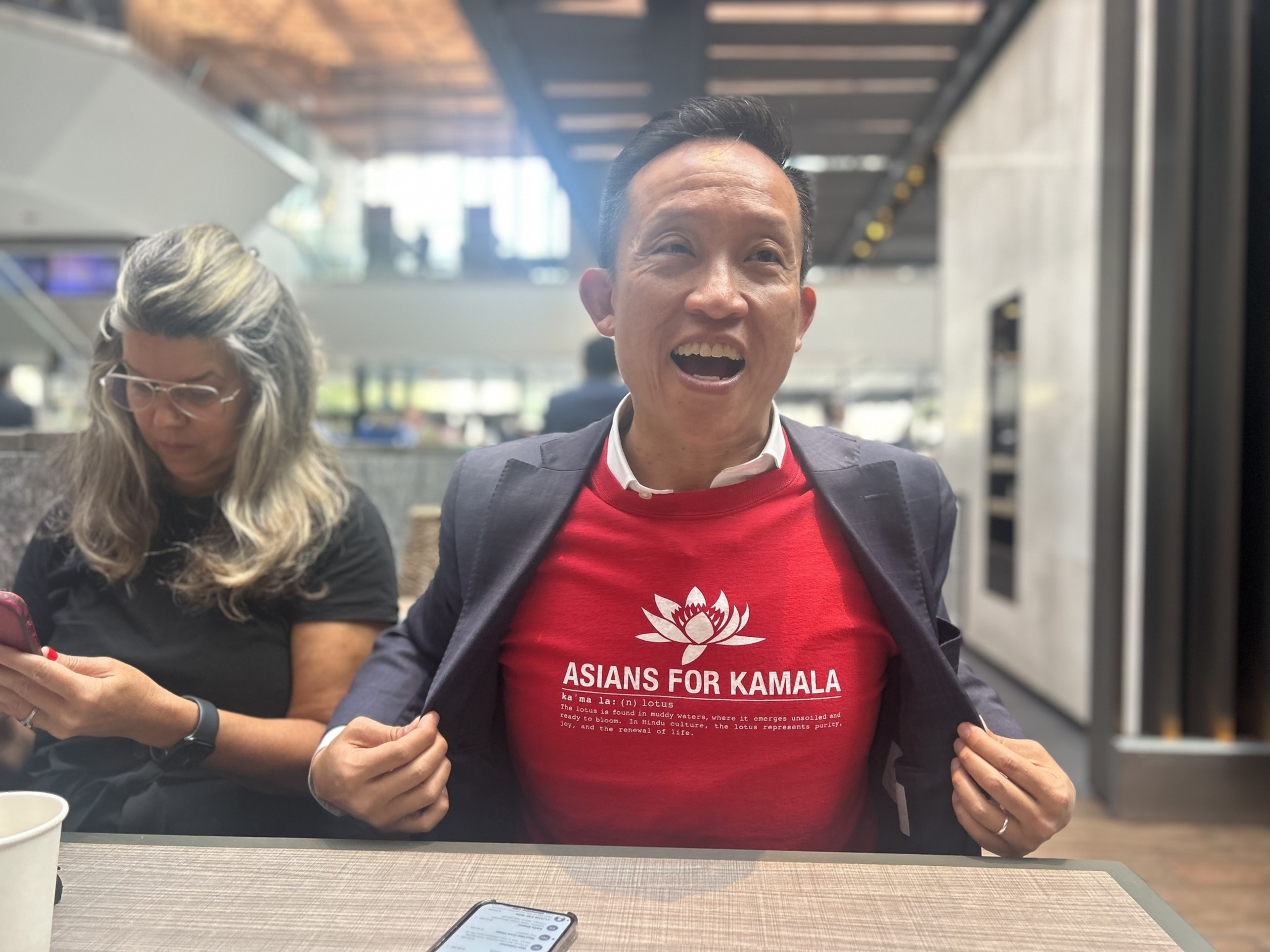 A smiling man opens his jacket to reveal a red shirt reading &quot;Asians for Kamala&quot; with a lotus logo. Beside him, a woman with glasses focuses on her phone.