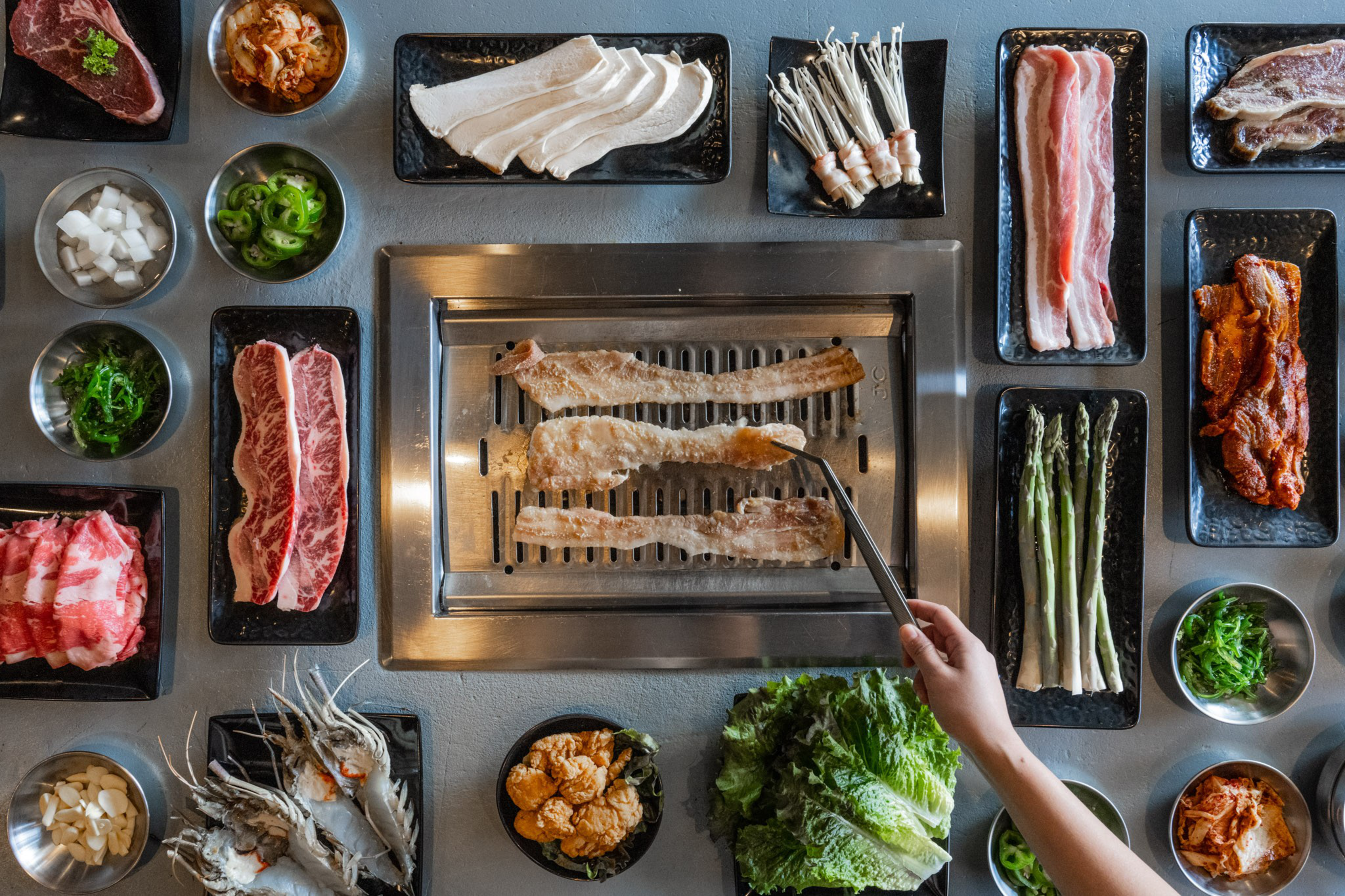 Various raw meats, seafood, mushrooms, vegetables, and greens surround a grill with several strips of meat being cooked, with a hand holding tongs.