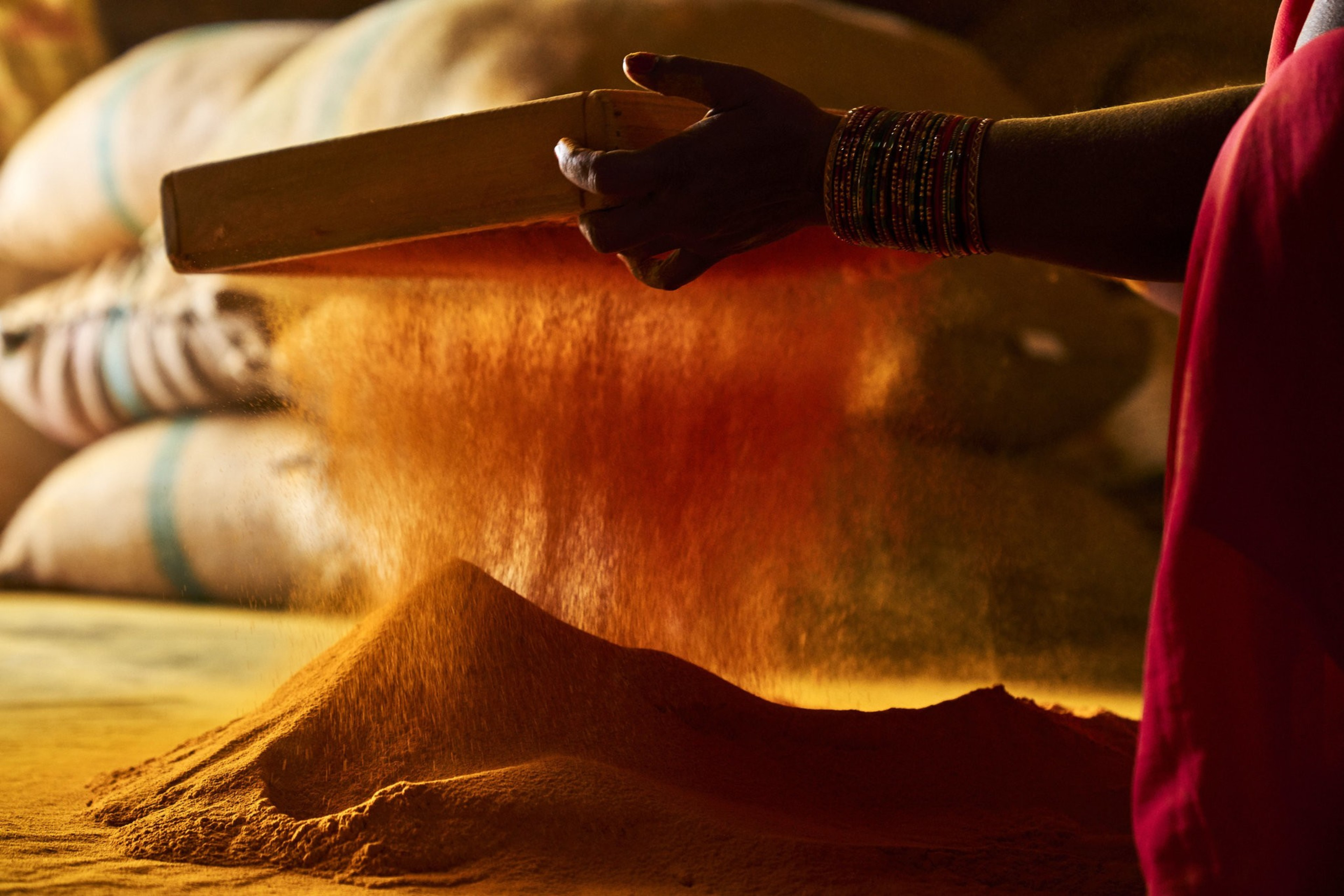 A person with decorated wrists sifts vibrant orange powder through a rectangular sieve, creating a glowing dust cloud over a pile on the ground.