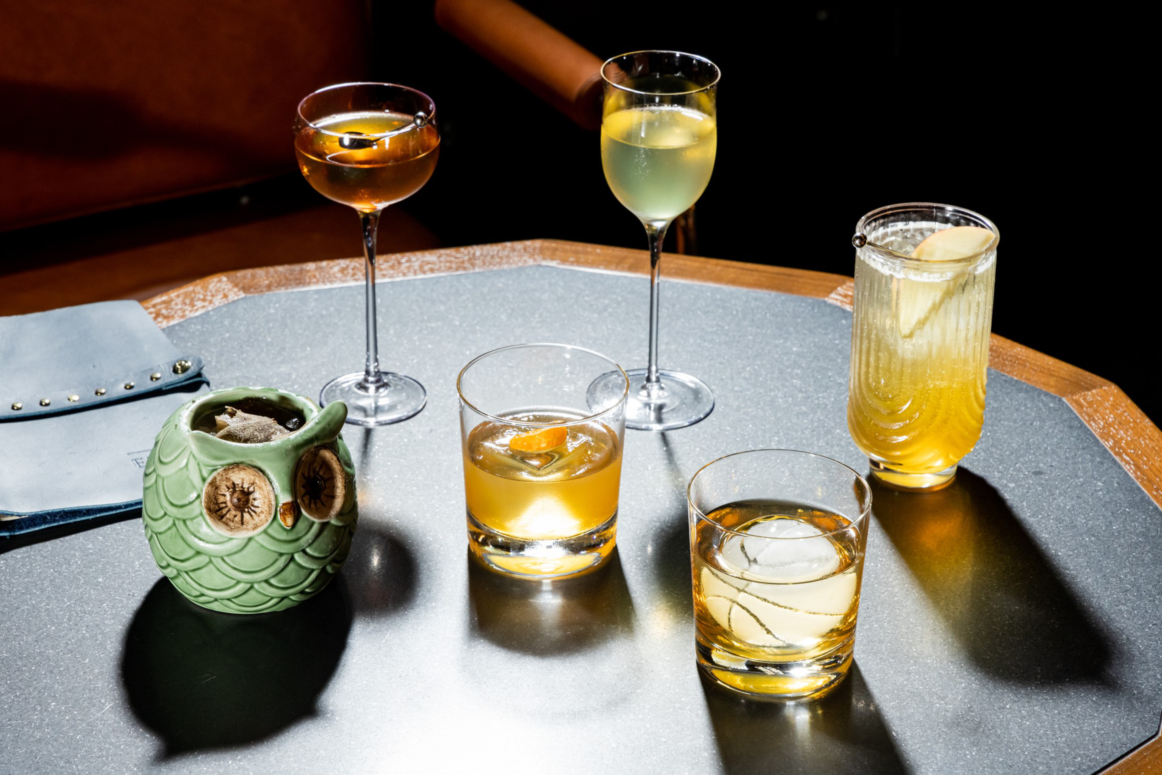 Five different cocktails in various glass styles sit on a table, including a green owl-shaped mug and a light blue clutch bag nearby.