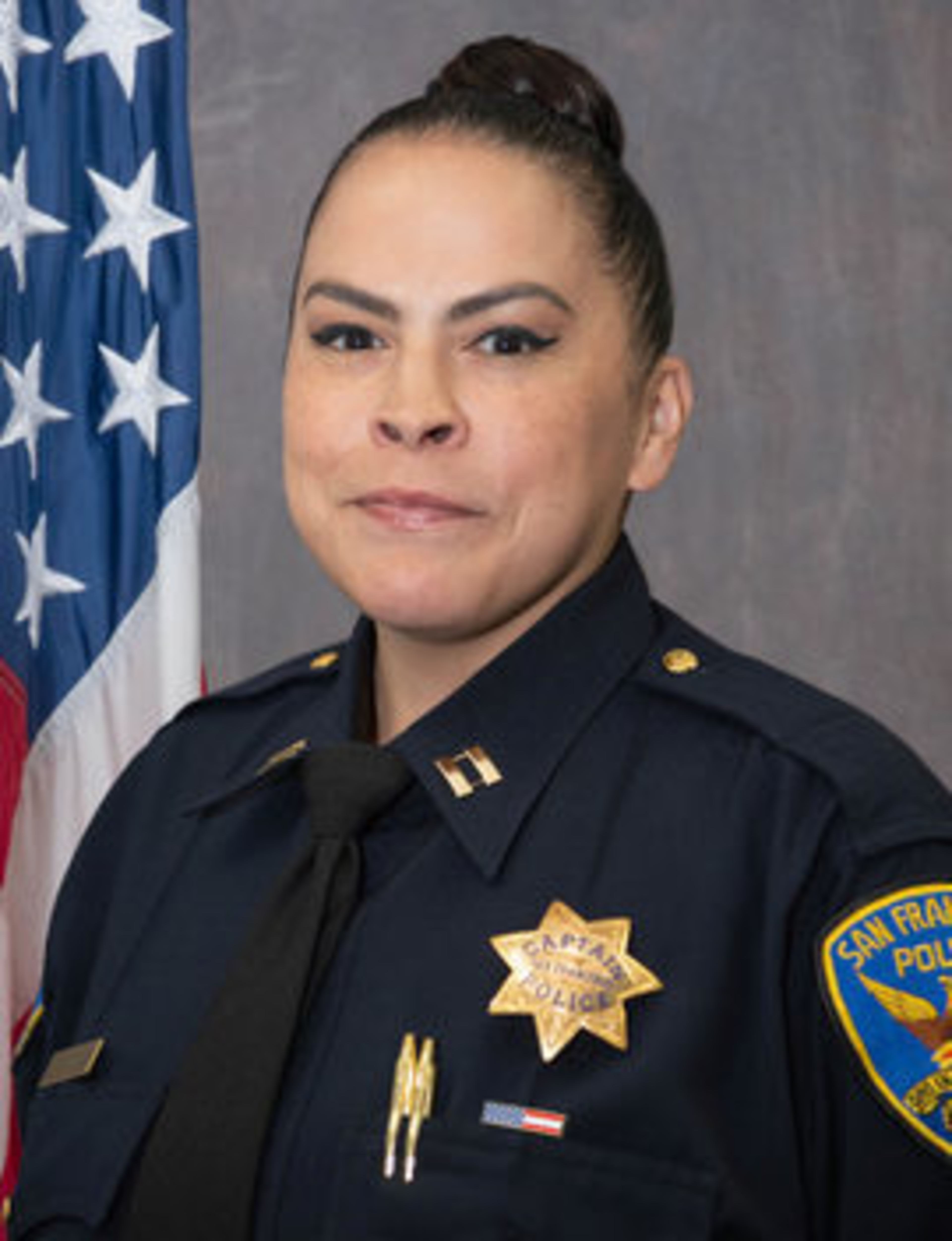 A female police officer in a San Francisco Police Department uniform with captain insignia stands in front of a U.S. flag and a gray background.