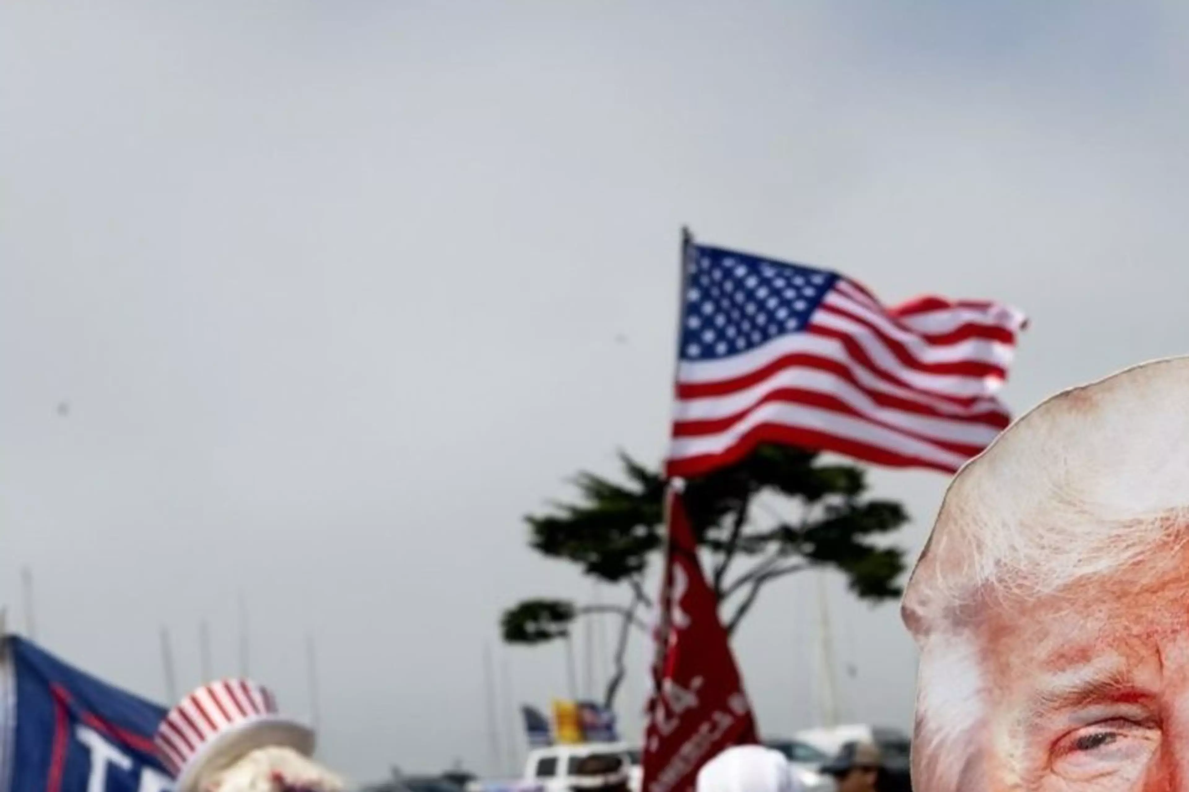 A person wearing a Donald Trump mask is flexing their arms in front of a group of people holding American flags and "Trump" banners. Other attendees are also visible.
