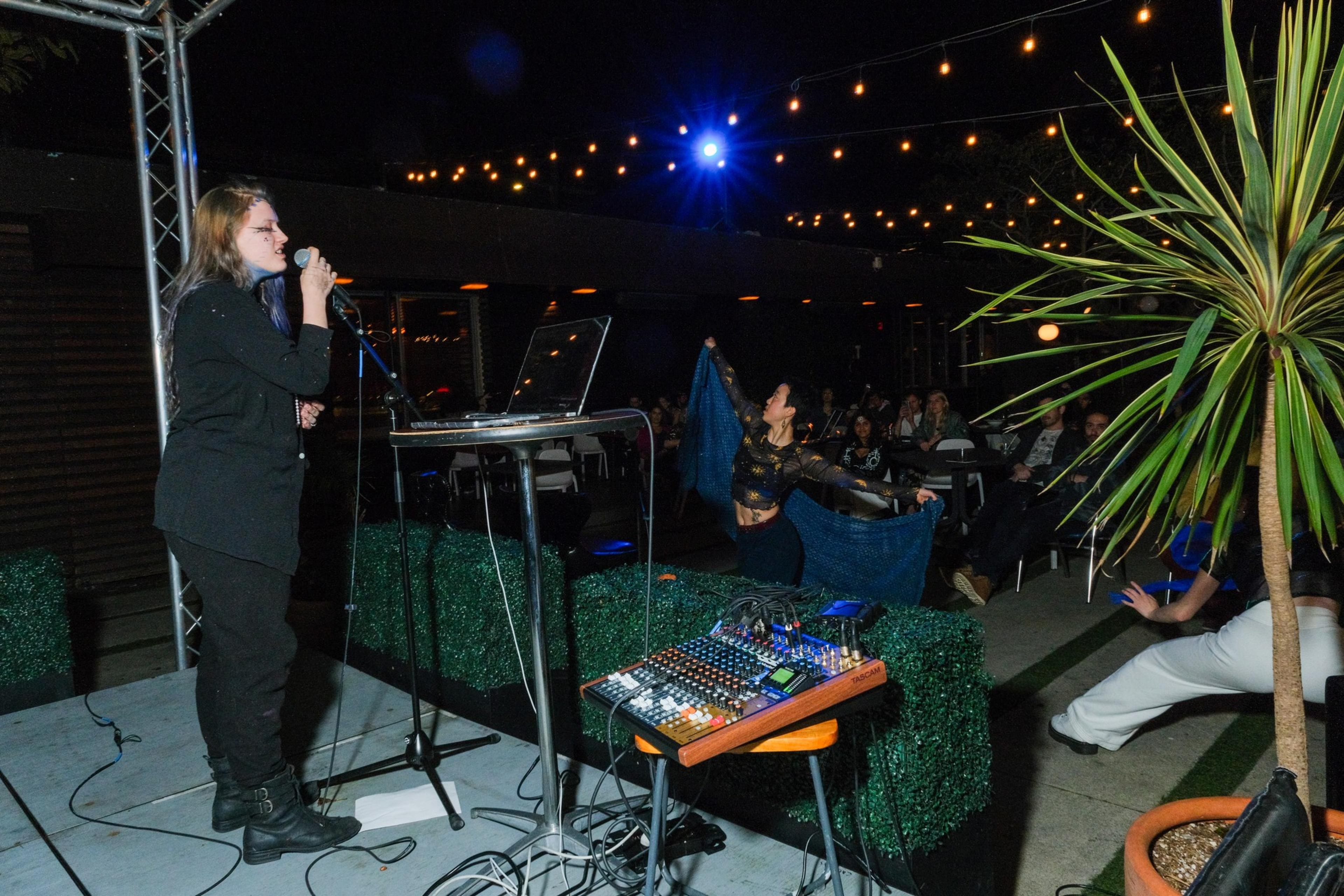 A singer performs on stage next to a dancer with flowing fabric, surrounded by an audience under string lights at night.