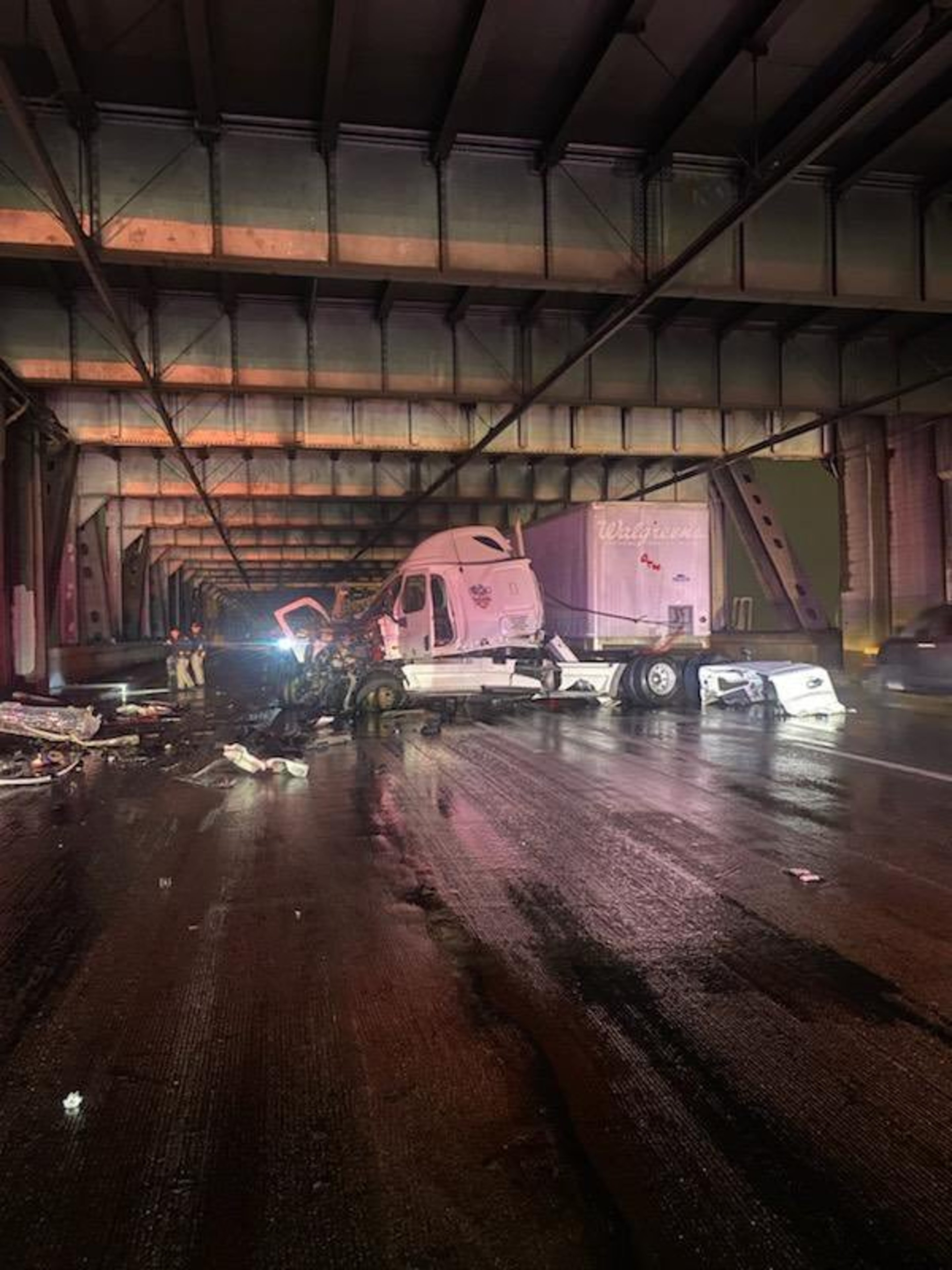 A damaged semi-truck is crashed under a bridge at night, debris scattered on the wet road. Emergency responders are present in the background.