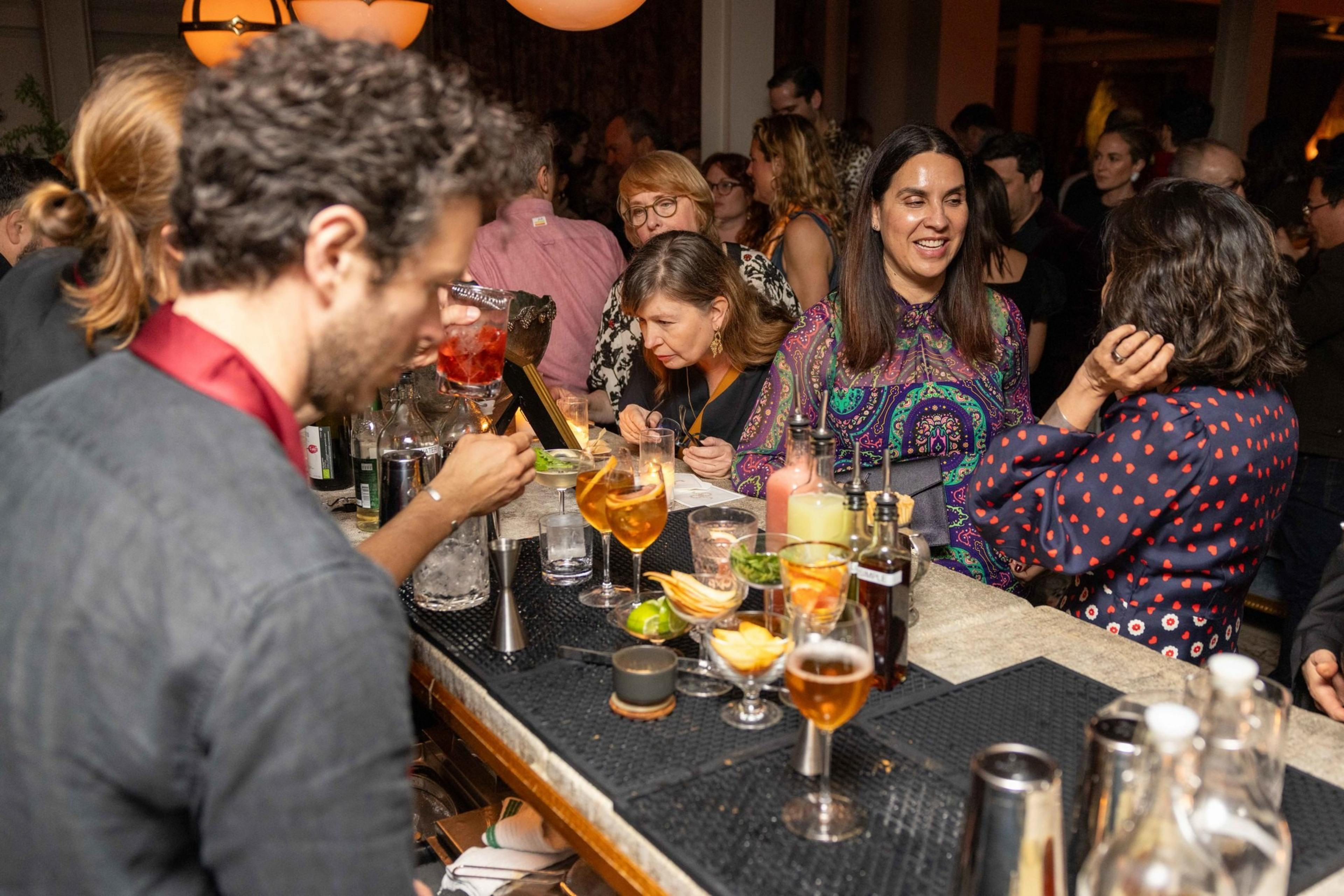 A crowded bar scene with a bartender mixing drinks and people socializing. Glasses, bottles, and garnishes are spread across the counter.