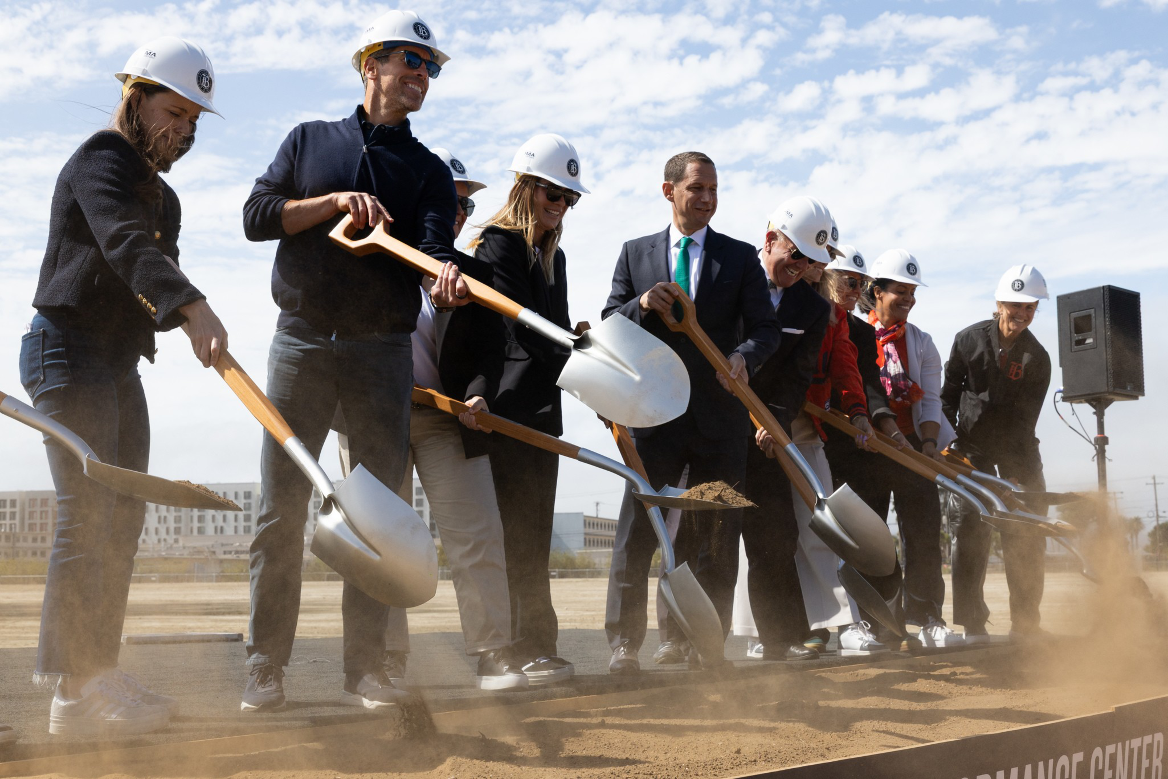 Eight people wearing white hard hats use shovels to dig into dirt at a groundbreaking ceremony on a sunny day.