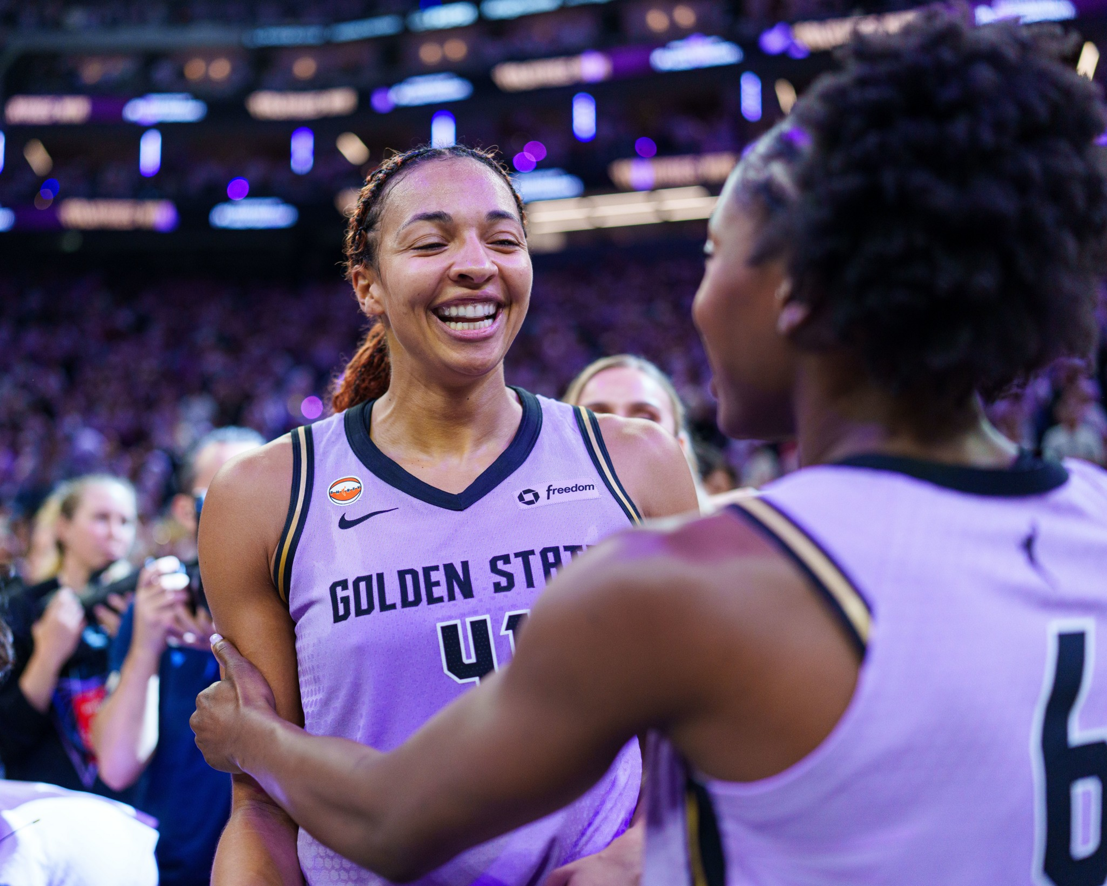 Two female basketball players in Golden State jerseys smile and embrace each other on the court with a cheering crowd in the background.