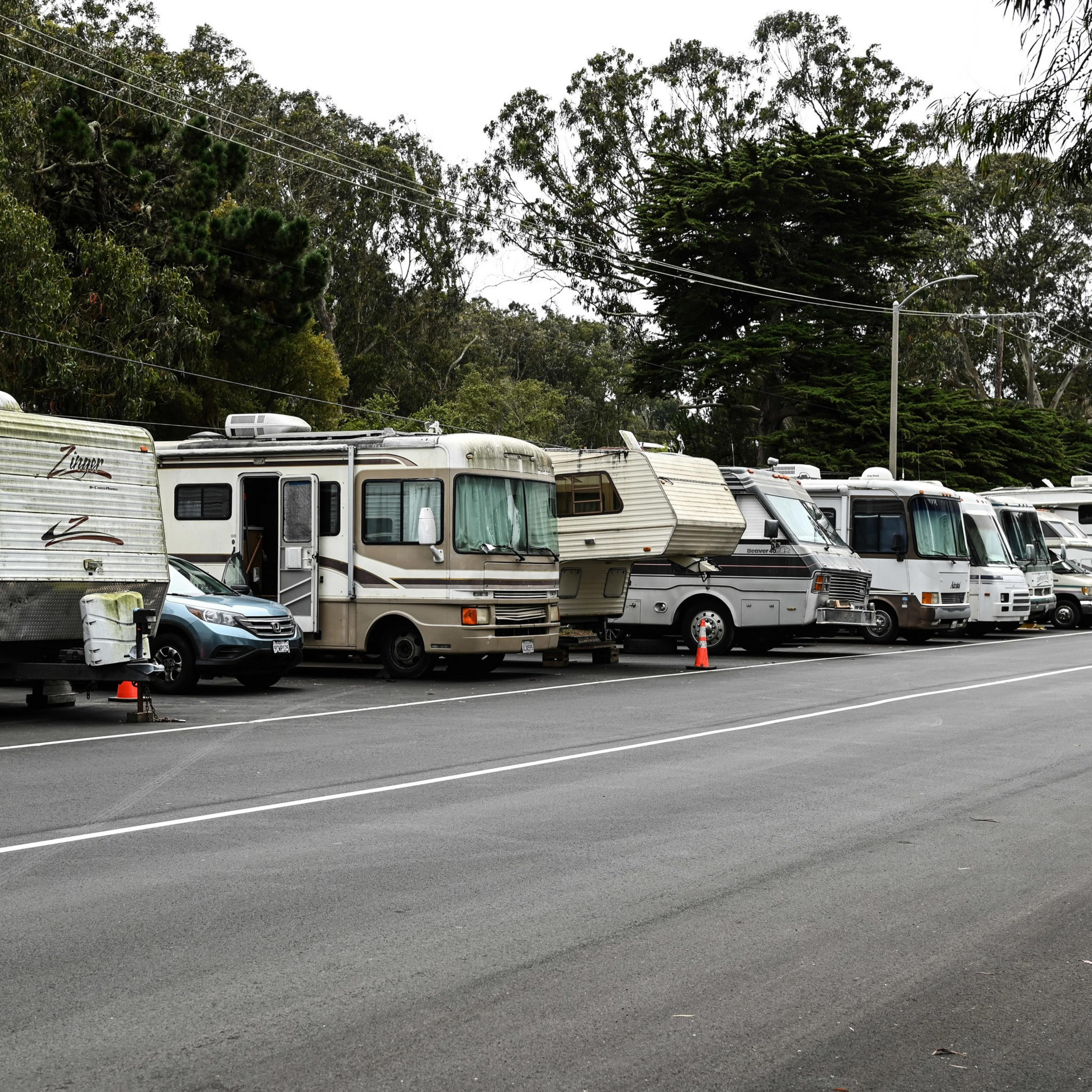 The image shows a row of parked RVs and trailers on a road, with trees in the background. It appears to be an RV park or a designated parking area for mobile homes.