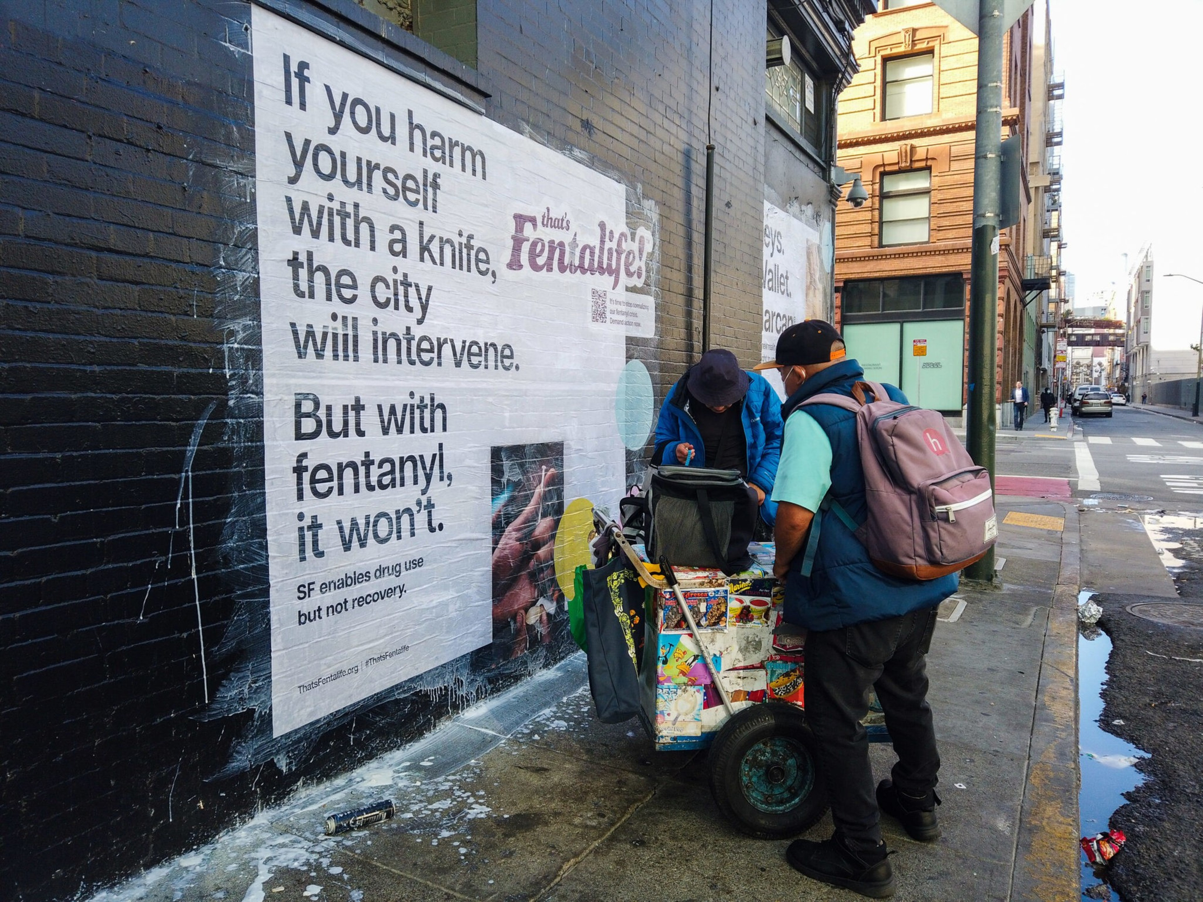 A large poster on a wall warns that the city intervenes if you harm yourself with a knife but not with fentanyl, while two people stand beside a cart covered in stickers.