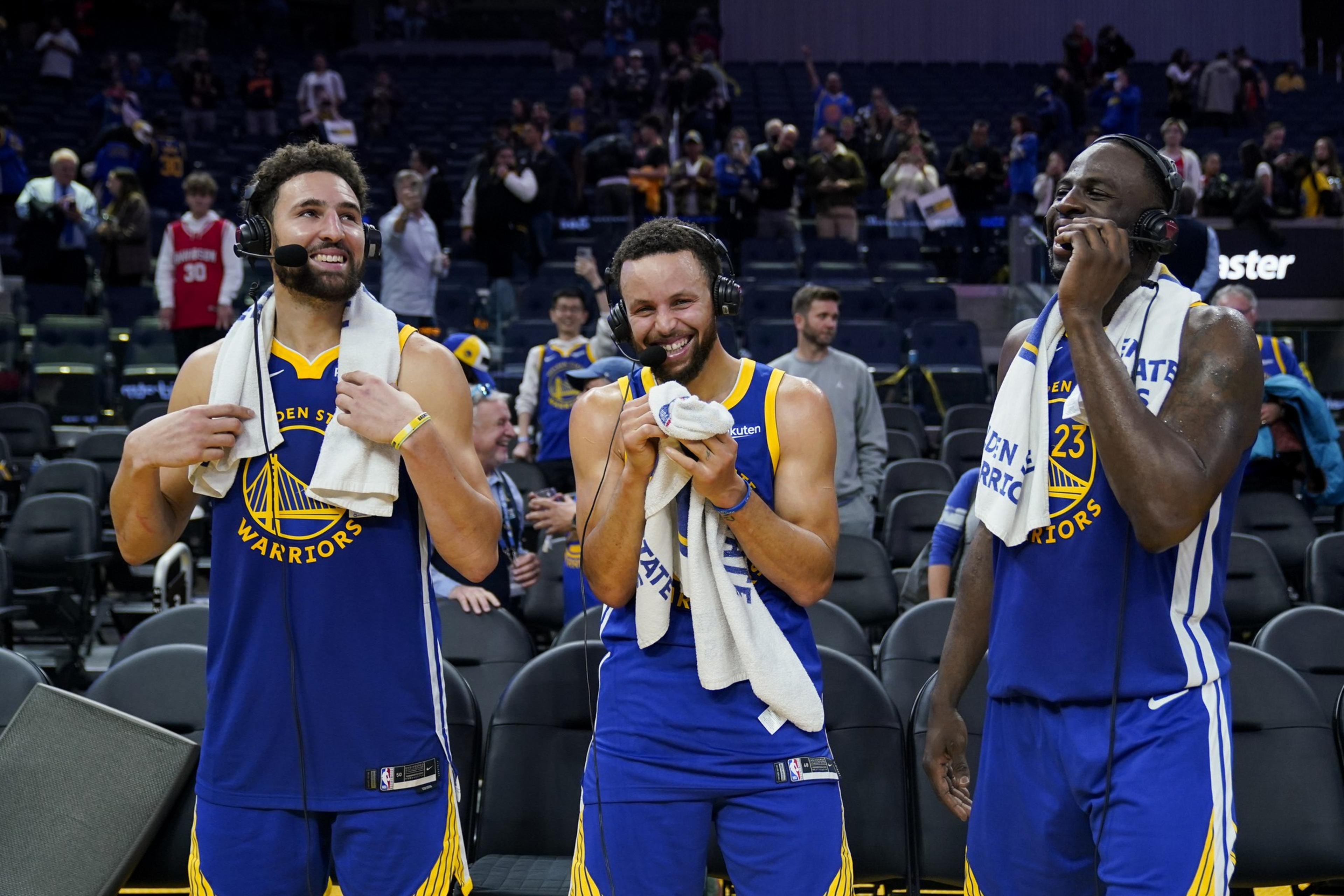 Three basketball players wearing Golden State Warriors jerseys stand smiling, holding towels and wearing headsets, with people in the background.