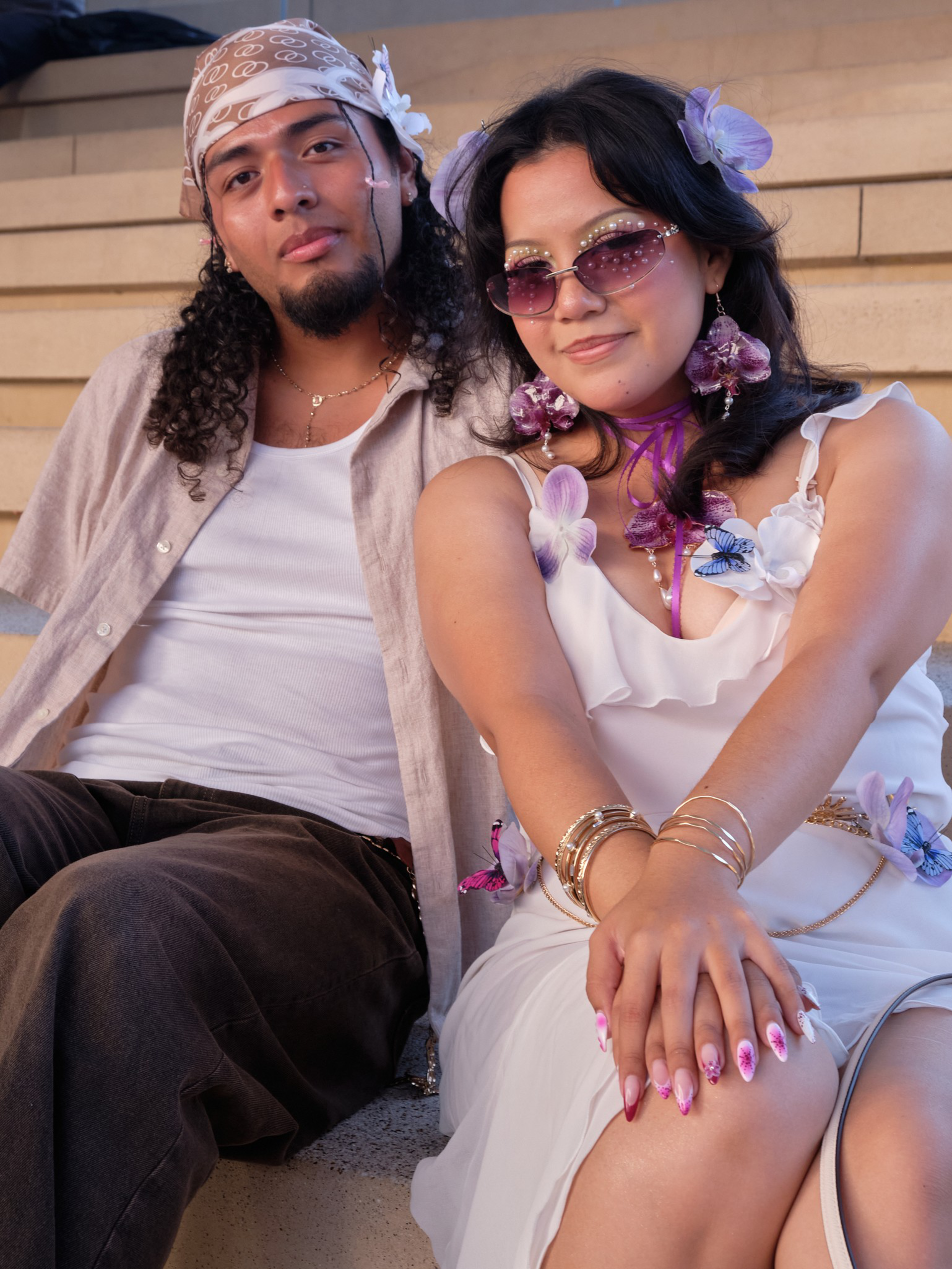 A man and woman sit closely on steps; he wears a bandana and casual clothes, she wears a white dress adorned with flowers, sunglasses, and butterfly accessories.