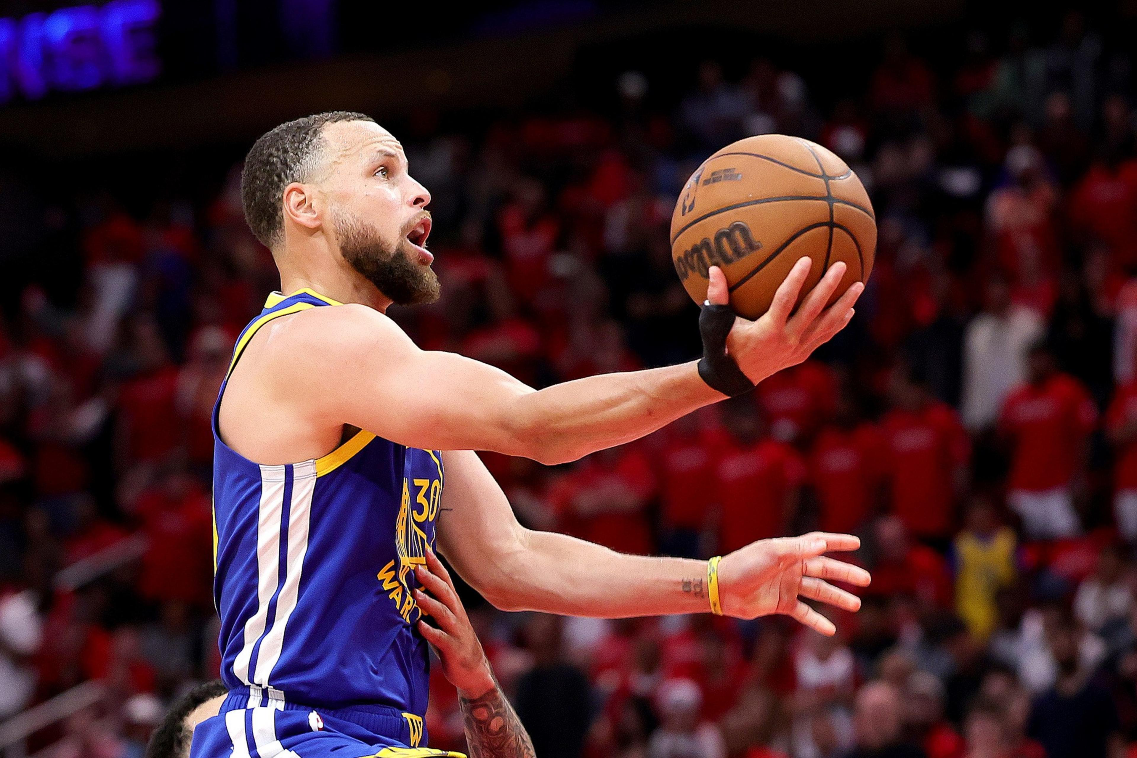 A basketball player in a blue and gold uniform is leaping with an outstretched arm, holding a basketball, against a crowd-filled background.