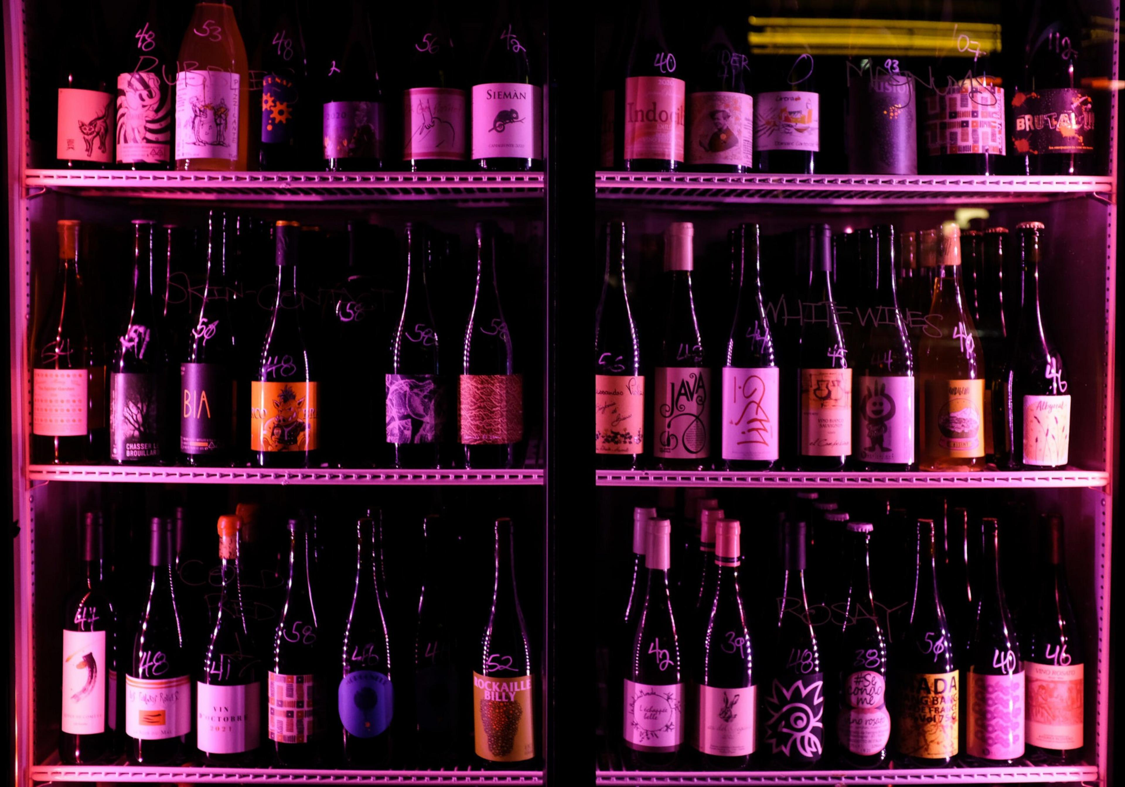 The image shows shelves of various wine bottles with unique labels, bathed in pinkish-purple lighting, arranged in a cooler or display case.