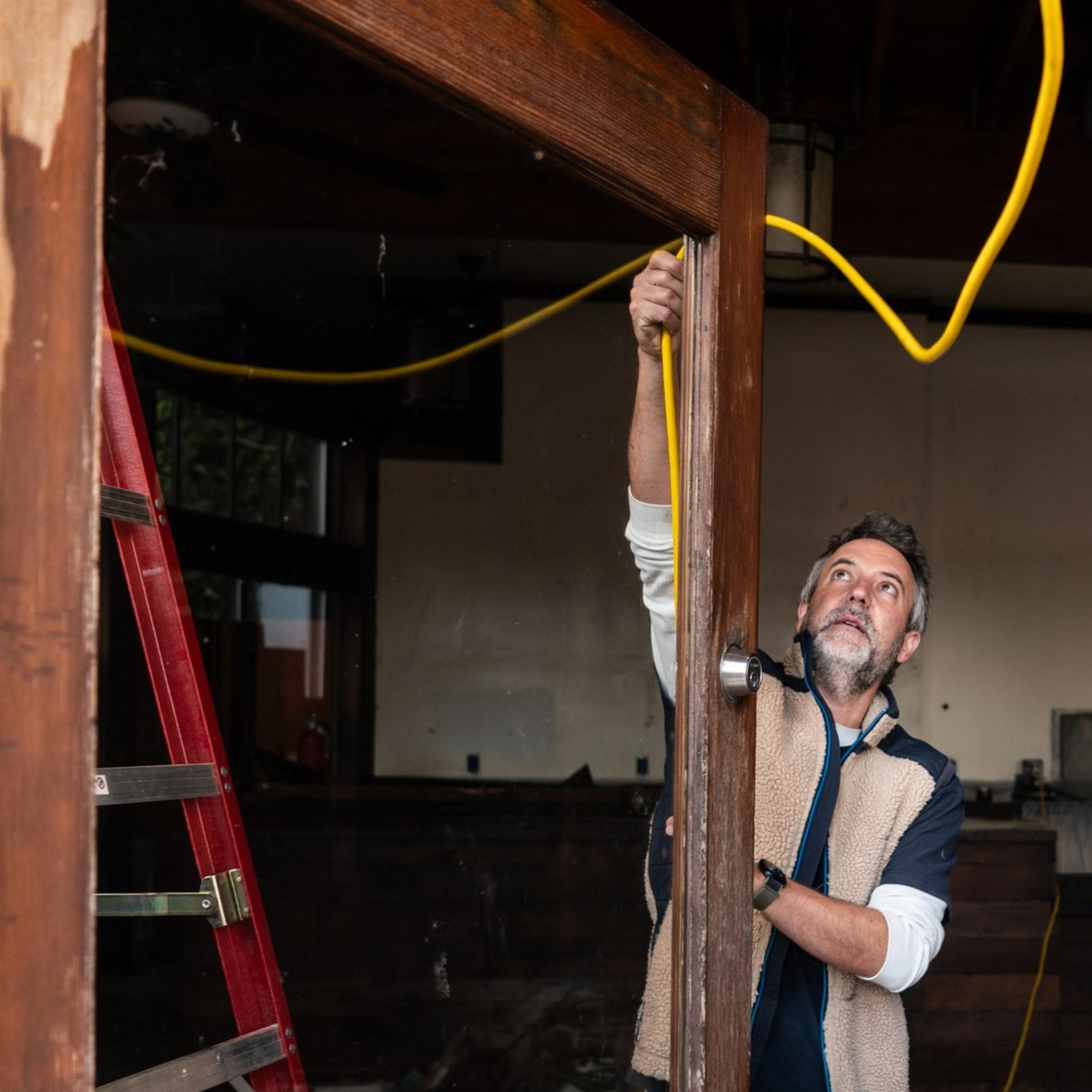A man is inspecting a wooden door frame inside a room under renovation, holding a yellow cable. A red ladder and plastic coverings are visible in the background.