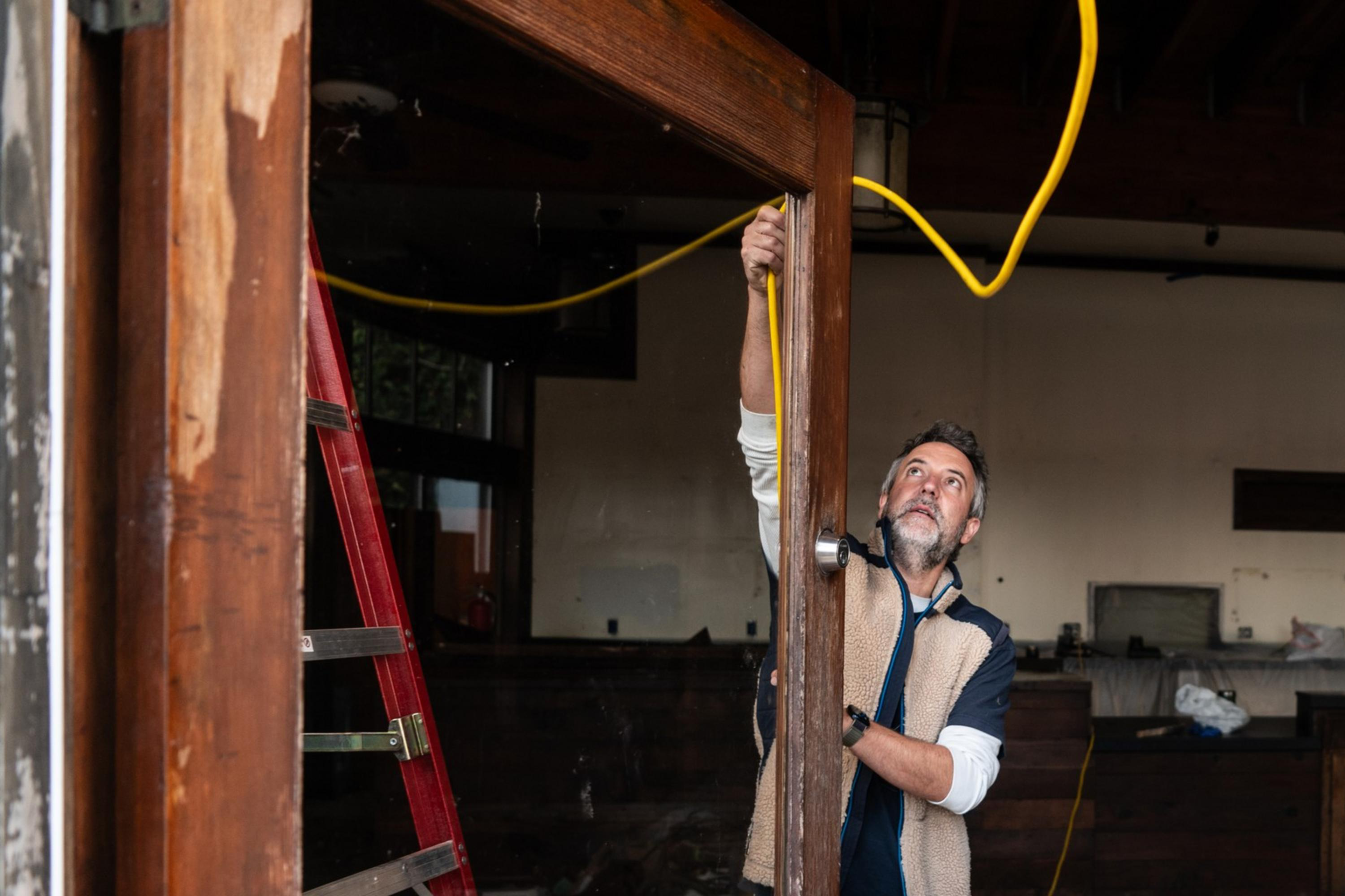 A man is inspecting a wooden door frame inside a room under renovation, holding a yellow cable. A red ladder and plastic coverings are visible in the background.