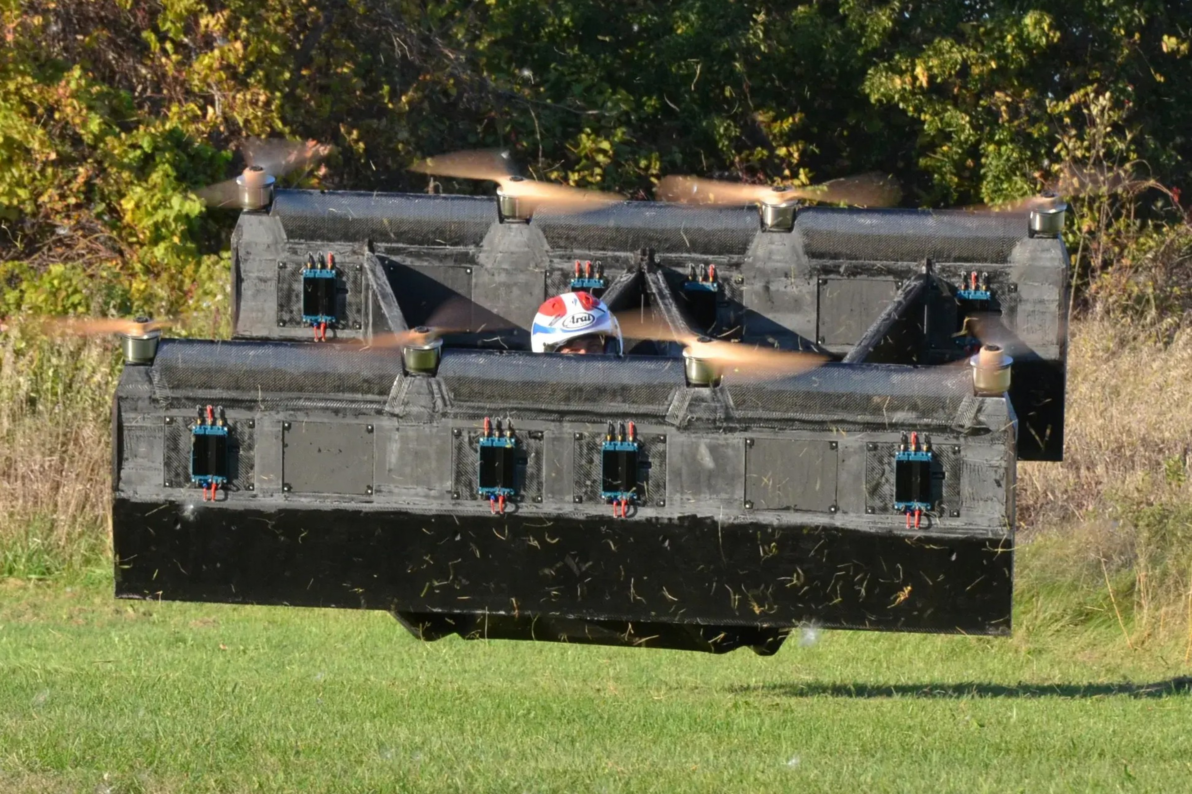 The image shows a flying machine resembling a large, black drone with eight rotating propellers, carrying a person wearing a helmet. It hovers low over grass with trees in the background.