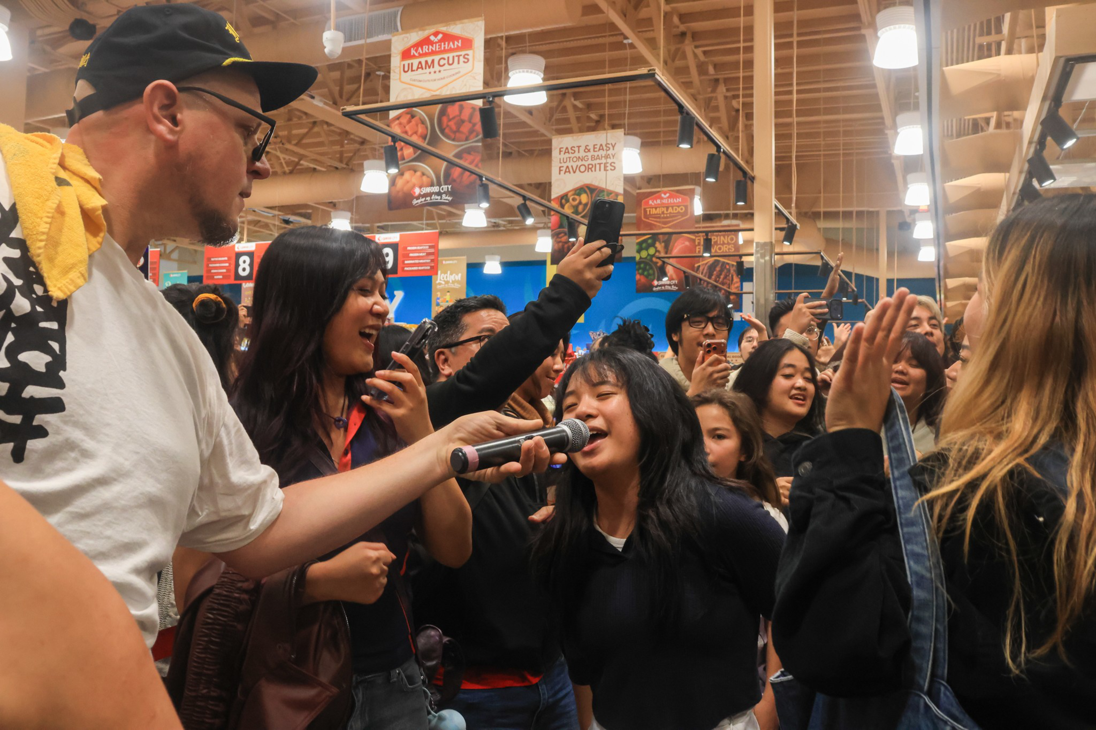 A man holds a microphone for a woman singing passionately while a crowd around them cheers, claps, and records the moment on their phones.