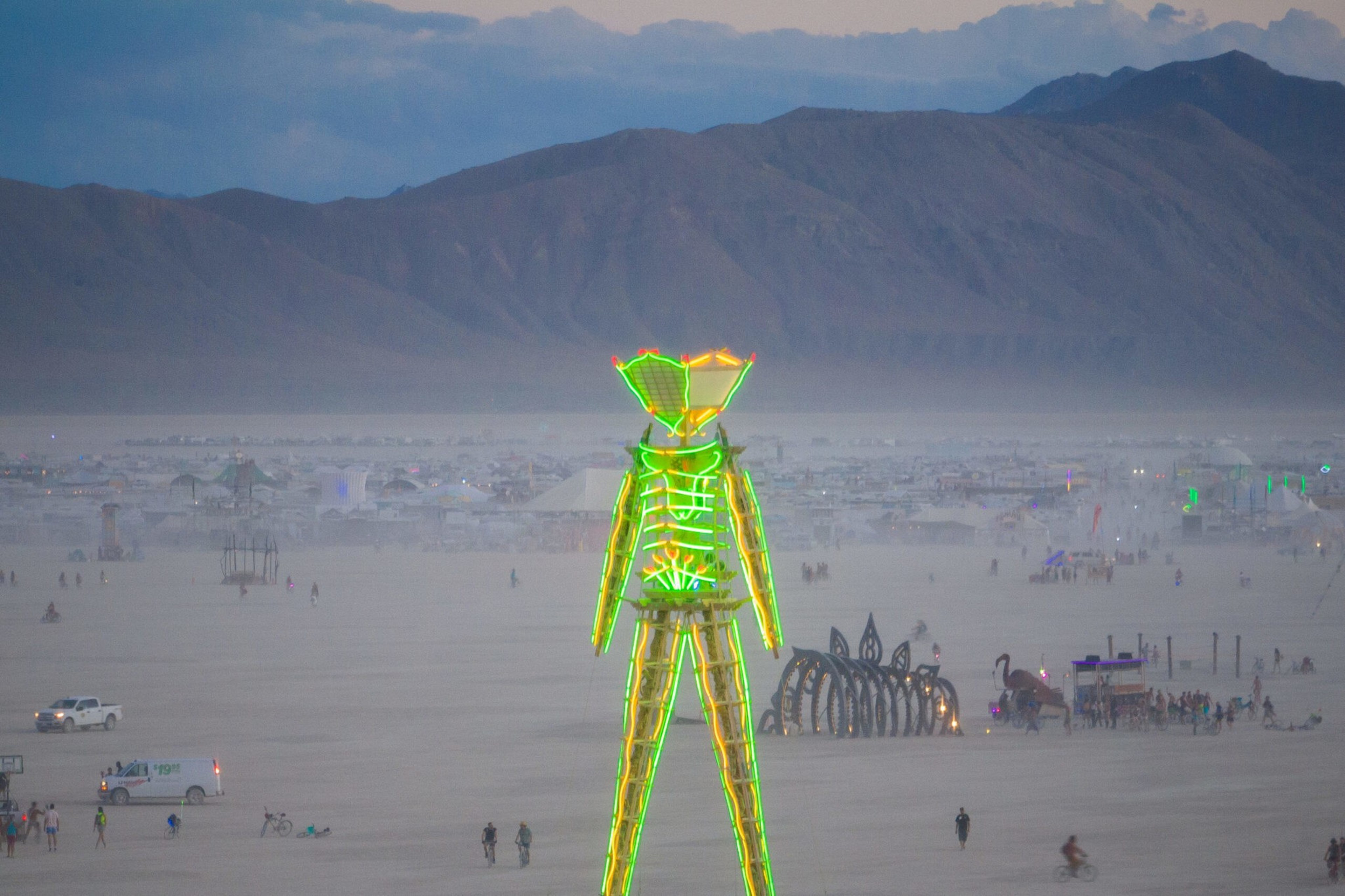 A large neon-lit humanoid figure stands on a desert plain with scattered people, bicycles, white vehicles, and mountains in the background.