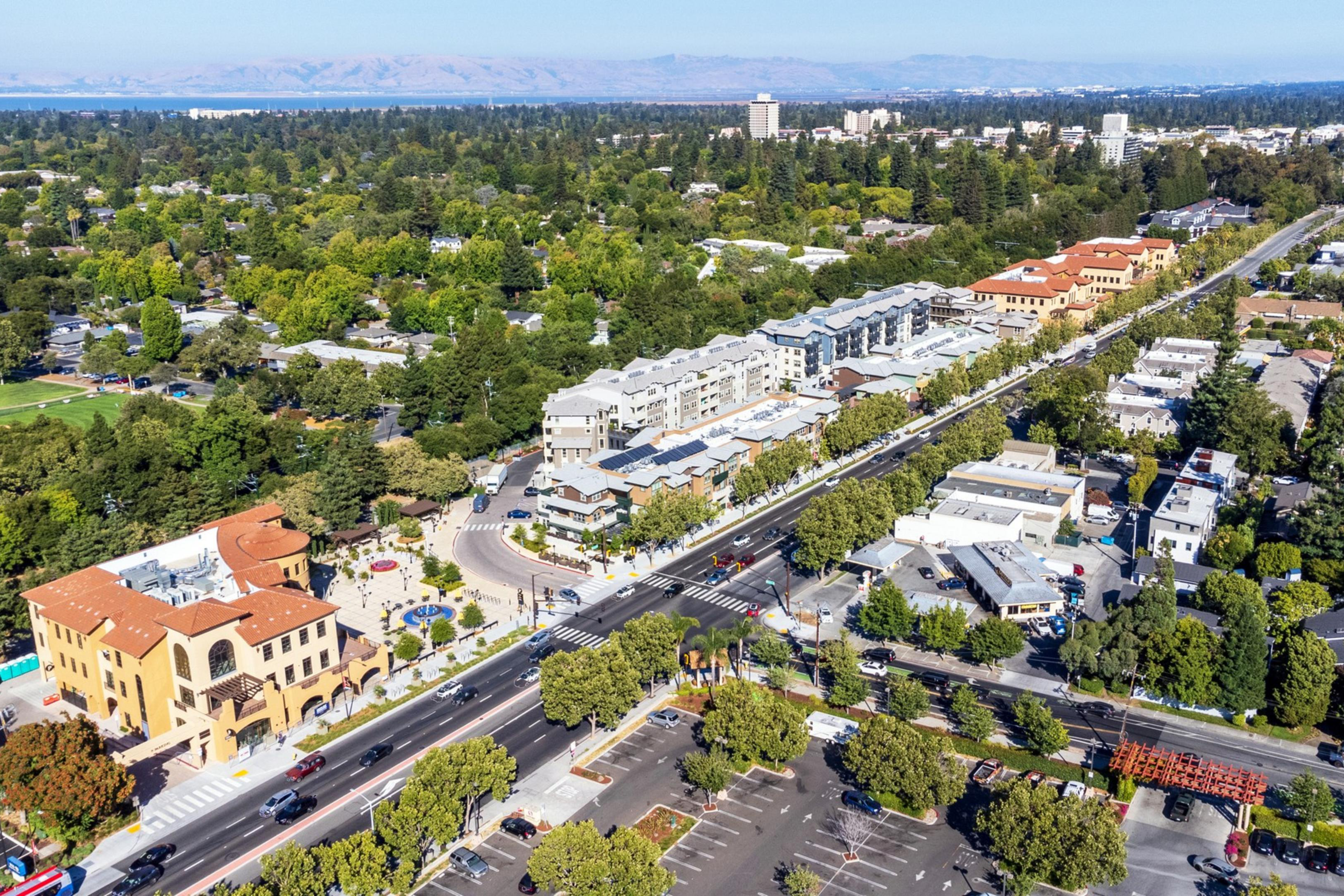 The image shows an aerial view of a suburban area with tree-lined streets, residential buildings, a baseball field, and some parking lots, under a clear sky.