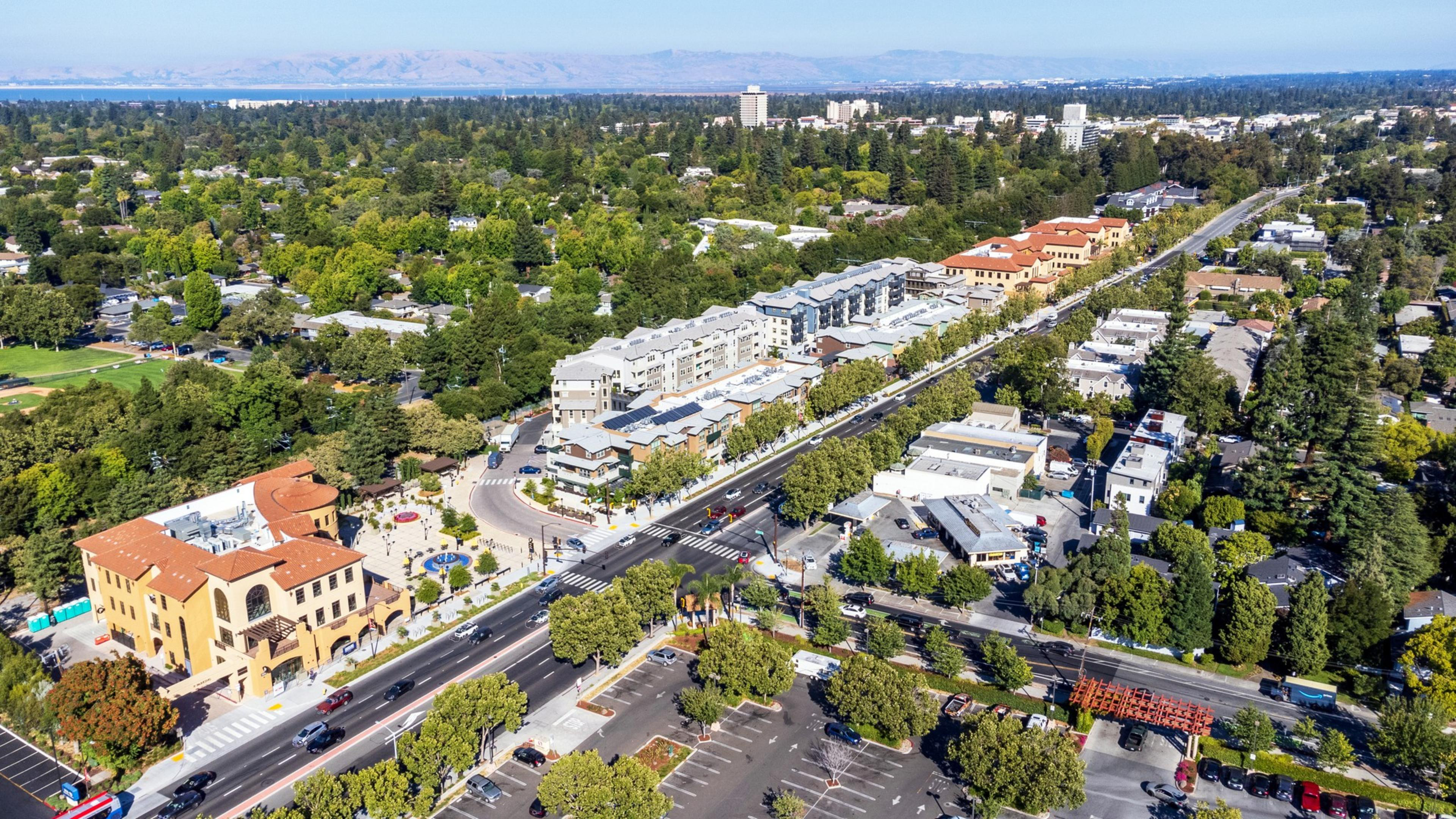 The image shows an aerial view of a suburban area with tree-lined streets, residential buildings, a baseball field, and some parking lots, under a clear sky.