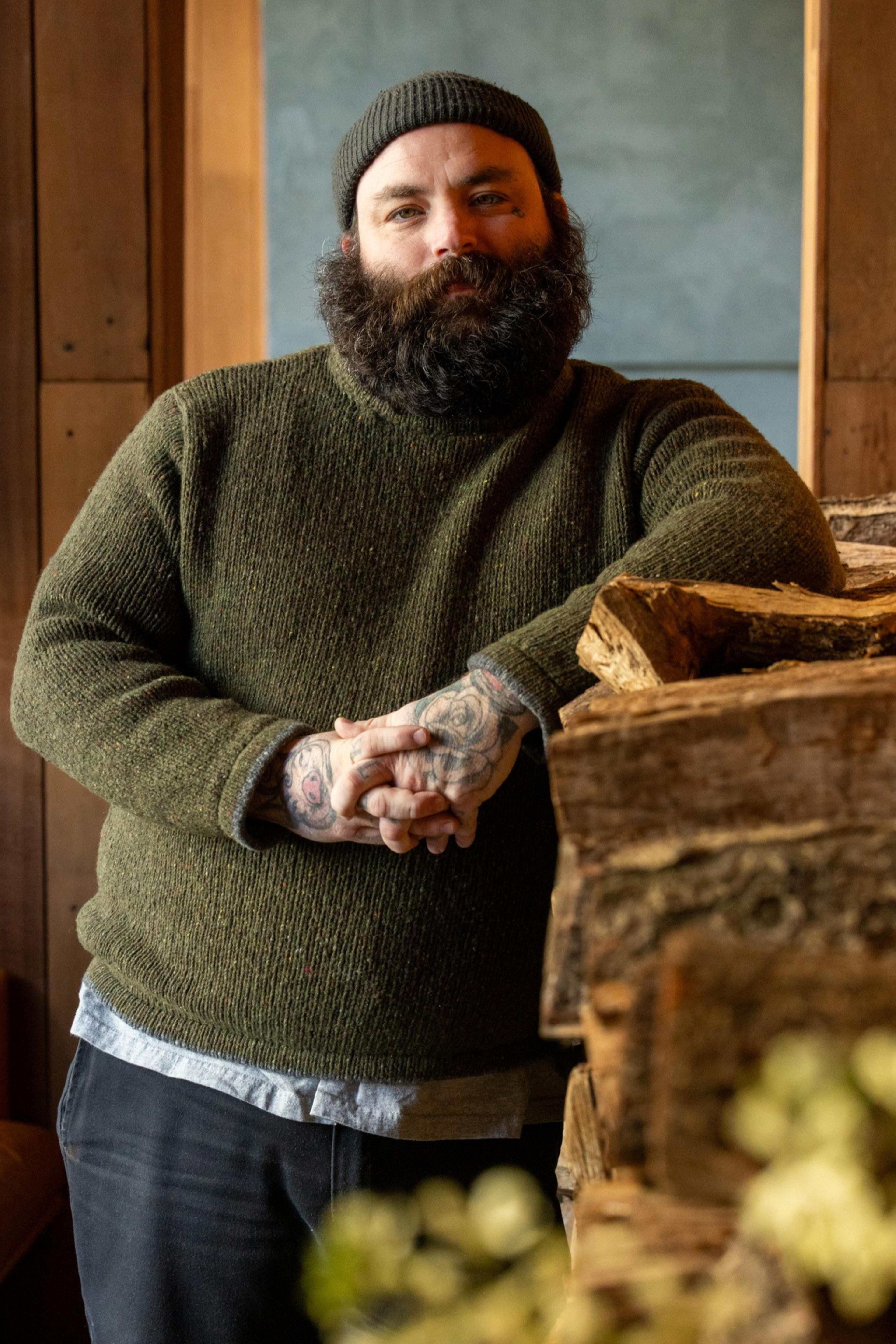 A bearded man in a green sweater and beanie is standing by a stack of firewood, with his tattooed hands clasped together.
