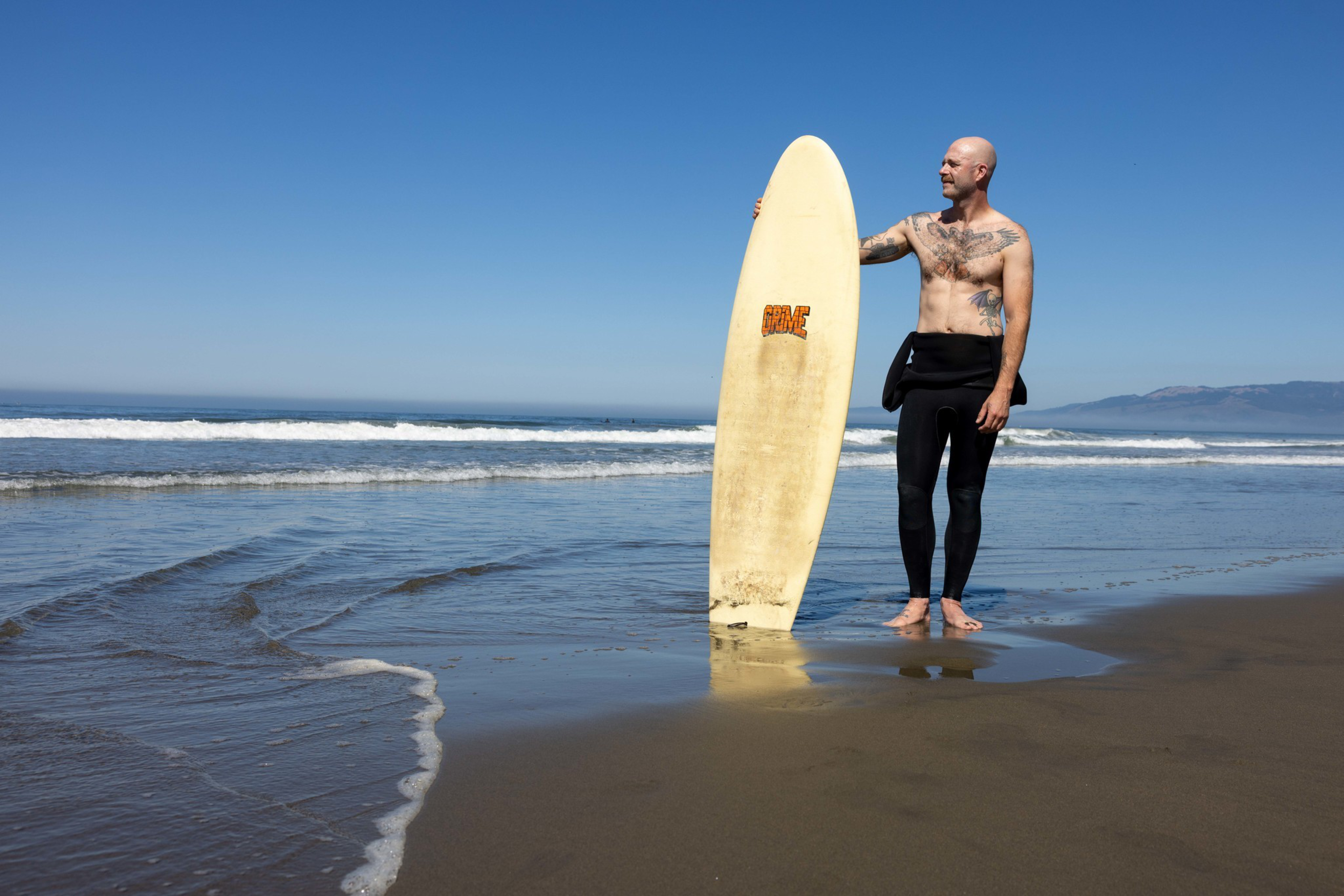A shirtless, tattooed man in black wetsuit pants stands barefoot on wet sand, holding a yellow surfboard near the ocean with waves and a clear blue sky.