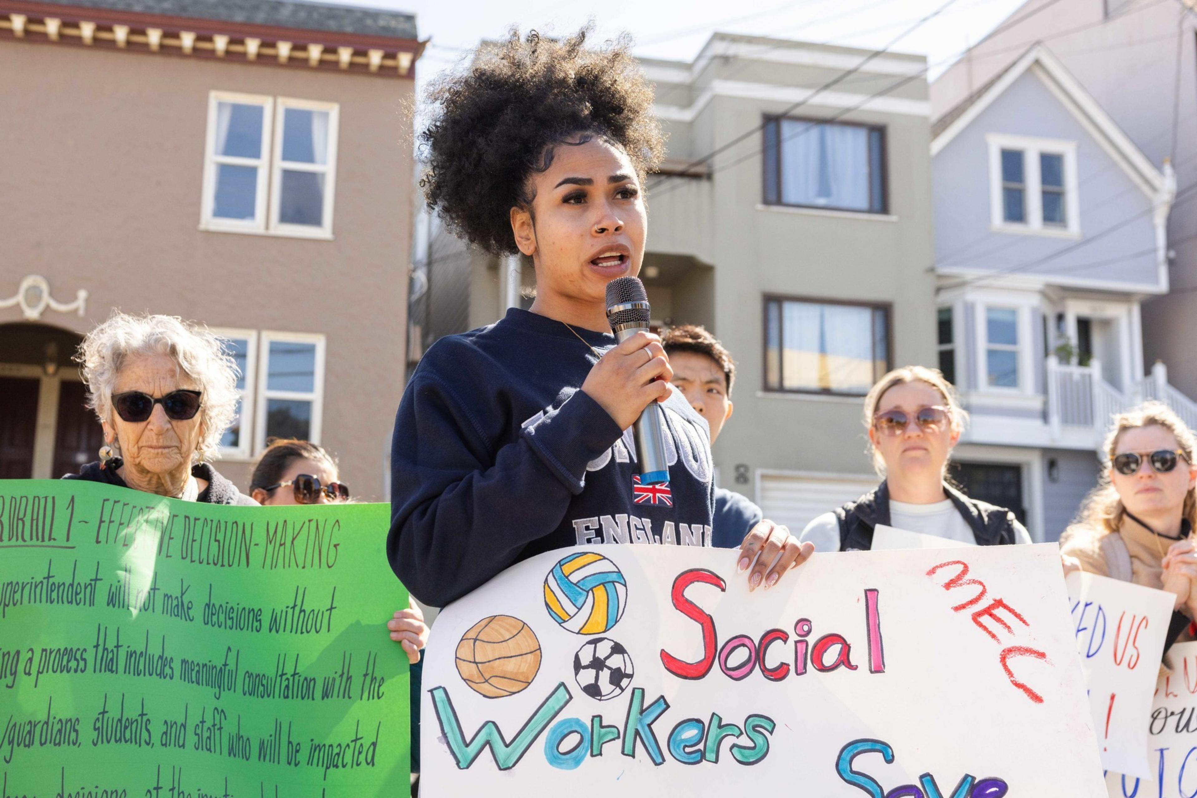 A person speaks into a microphone at a rally, holding a sign about social workers. Others stand nearby with signs, and houses are visible in the background.