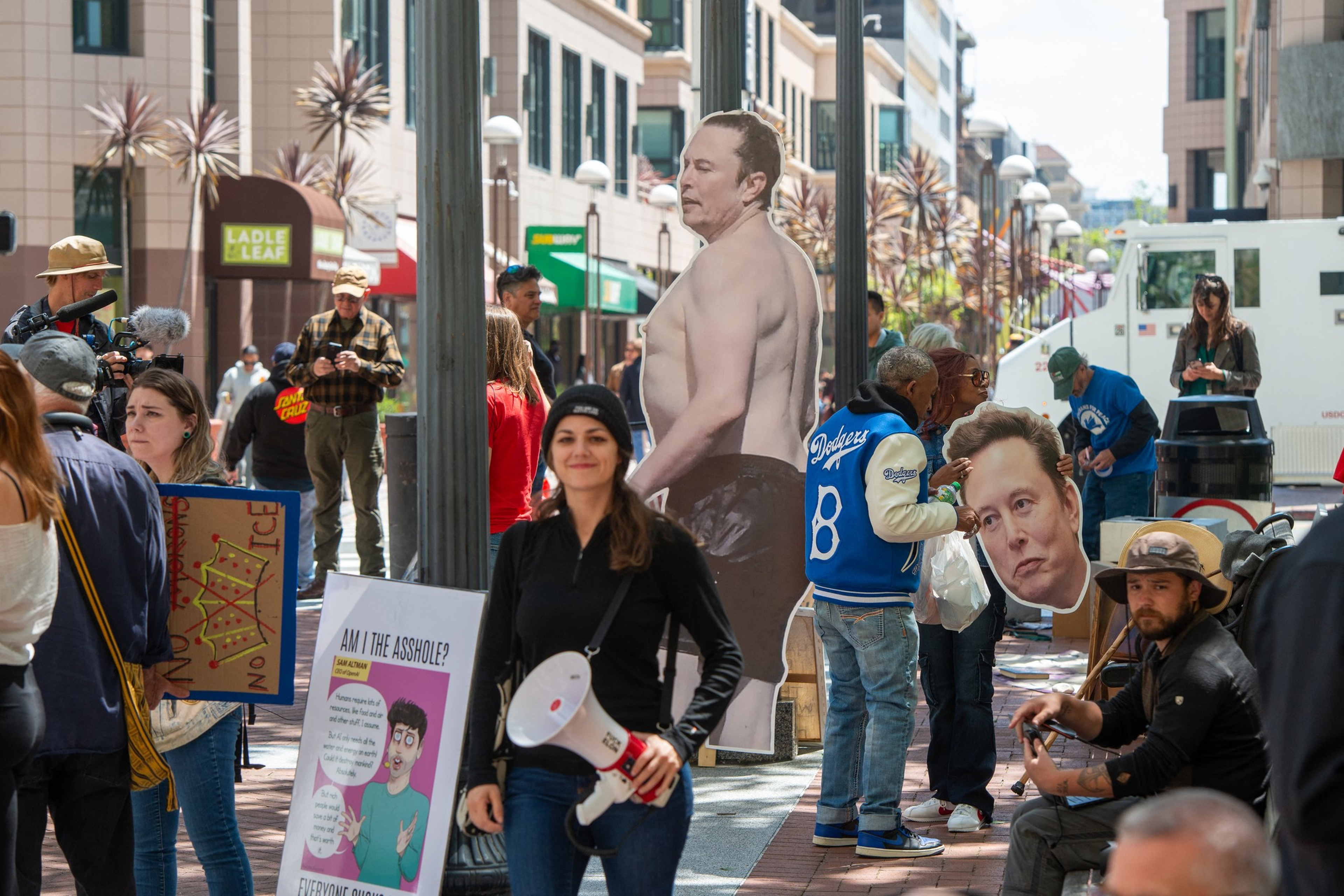 A street scene with people holding signs and a large cutout of a shirtless man, accompanied by a person holding a large mask of the same man’s face.