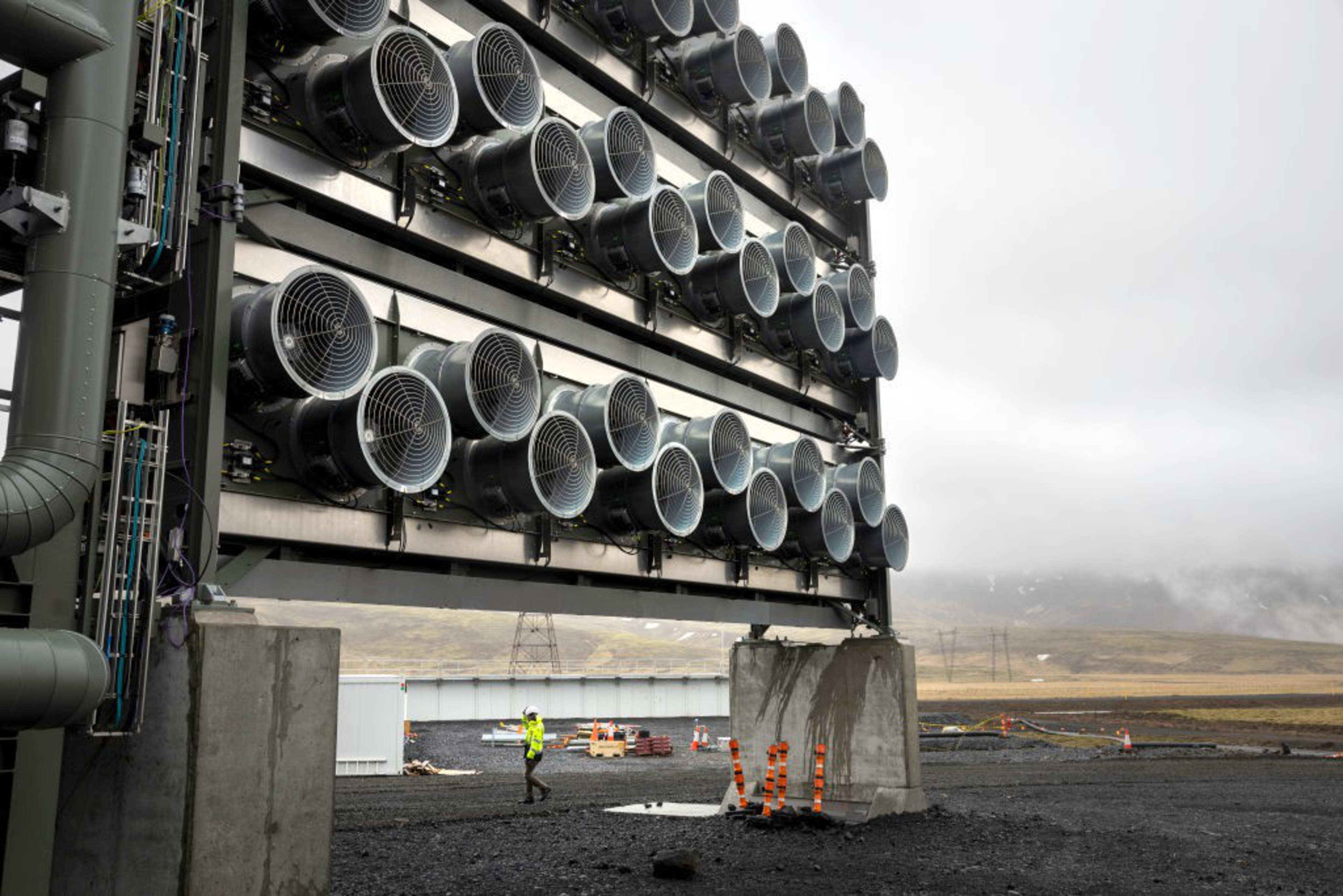 A large structure with rows of industrial fans stands amid a gray, misty landscape. A person in a high-visibility jacket walks below it on dark gravel.