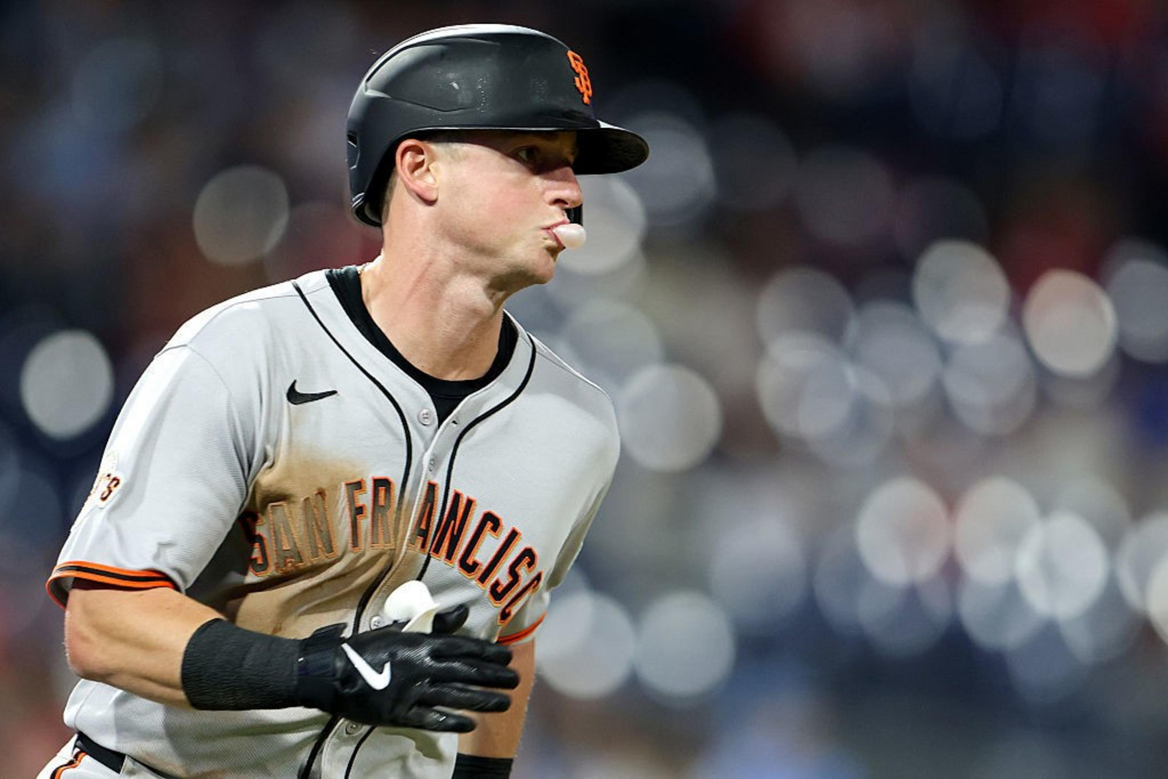 A baseball player in a gray uniform and black helmet runs with a focused expression, a bubblegum bubble emerging from his mouth. The background is a blur of lights.